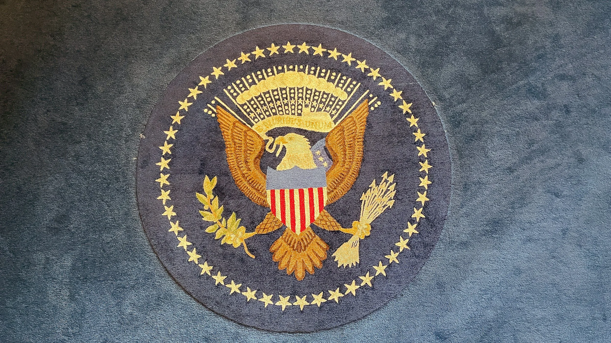 Close-up of an embroidered U.S. presidential seal on dark fabric, featuring an eagle, shield, olive branch, and arrows, surrounded by stars.