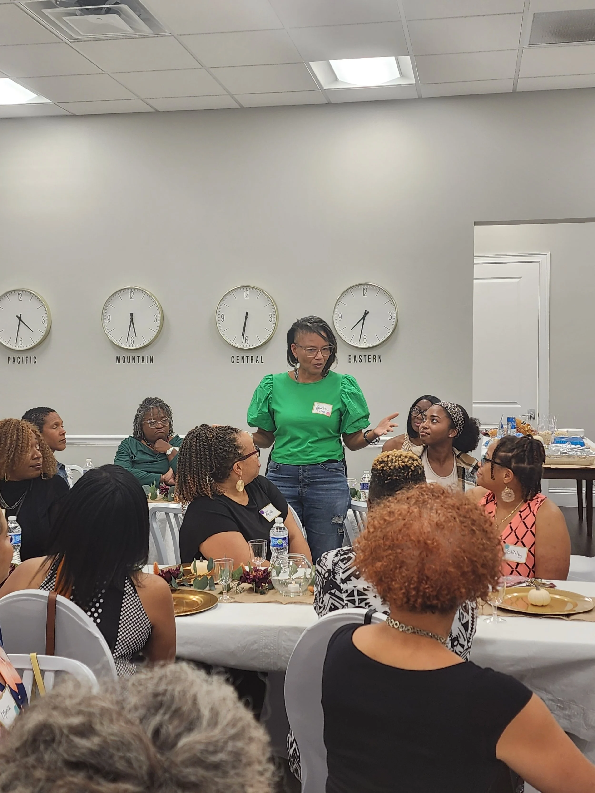 A woman in a green top speaking at a roundtable with several women seated, in a room with four clocks on the wall labeled Pacific, Mountain, Central, and Eastern, and a white door.