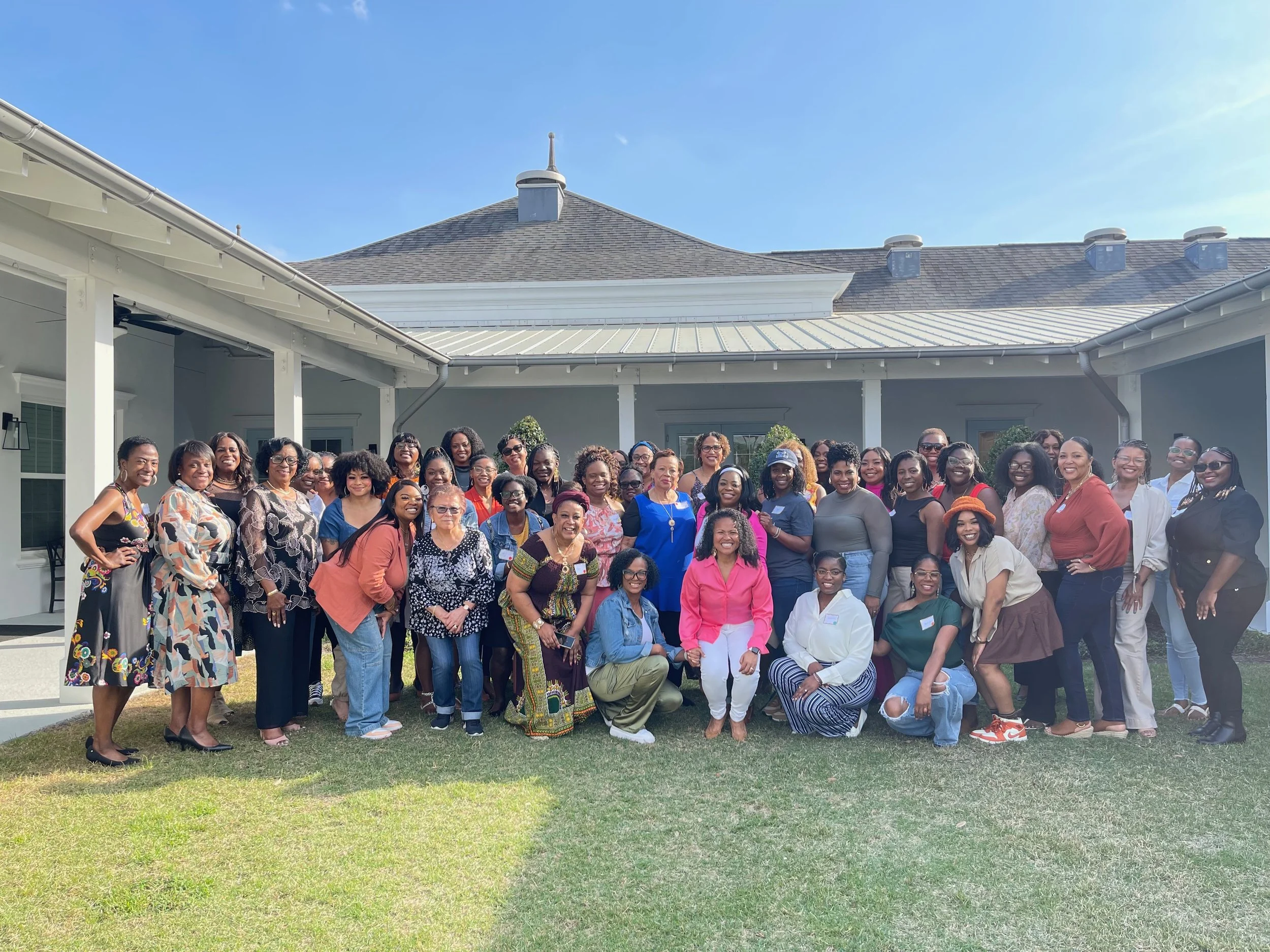 A large group of diverse women and a few men posing outdoors in front of a house under a blue sky, smiling and dressed in colorful clothing.