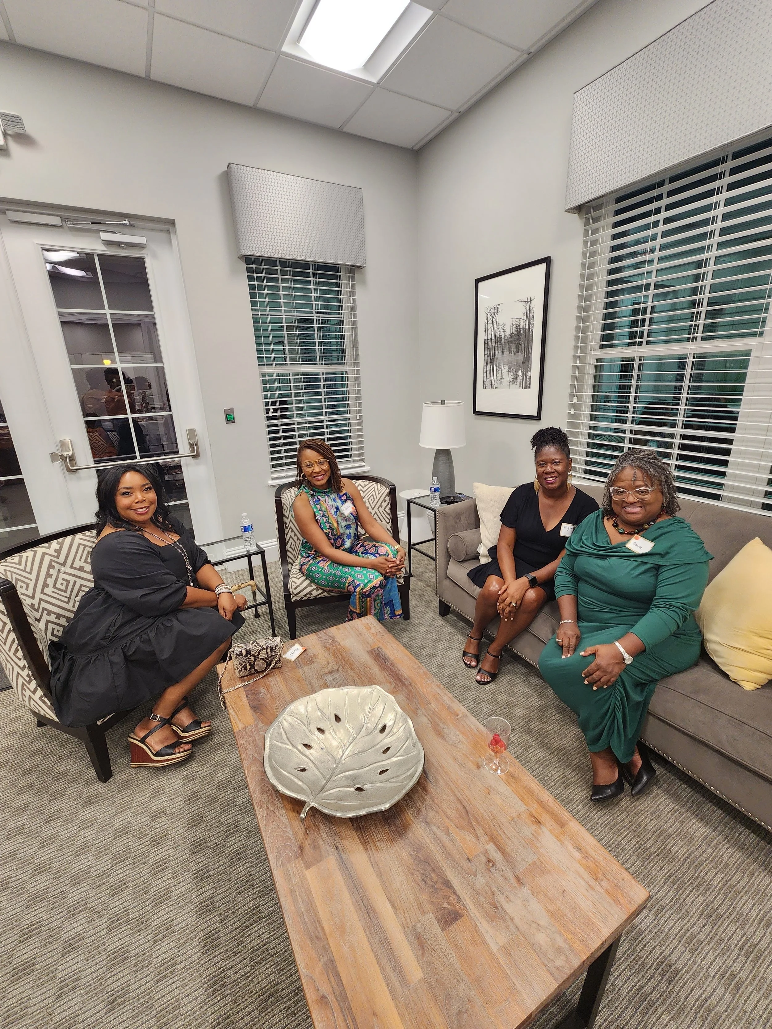 Five women sitting in a room with large windows, white walls, and modern furniture, smiling at the camera.