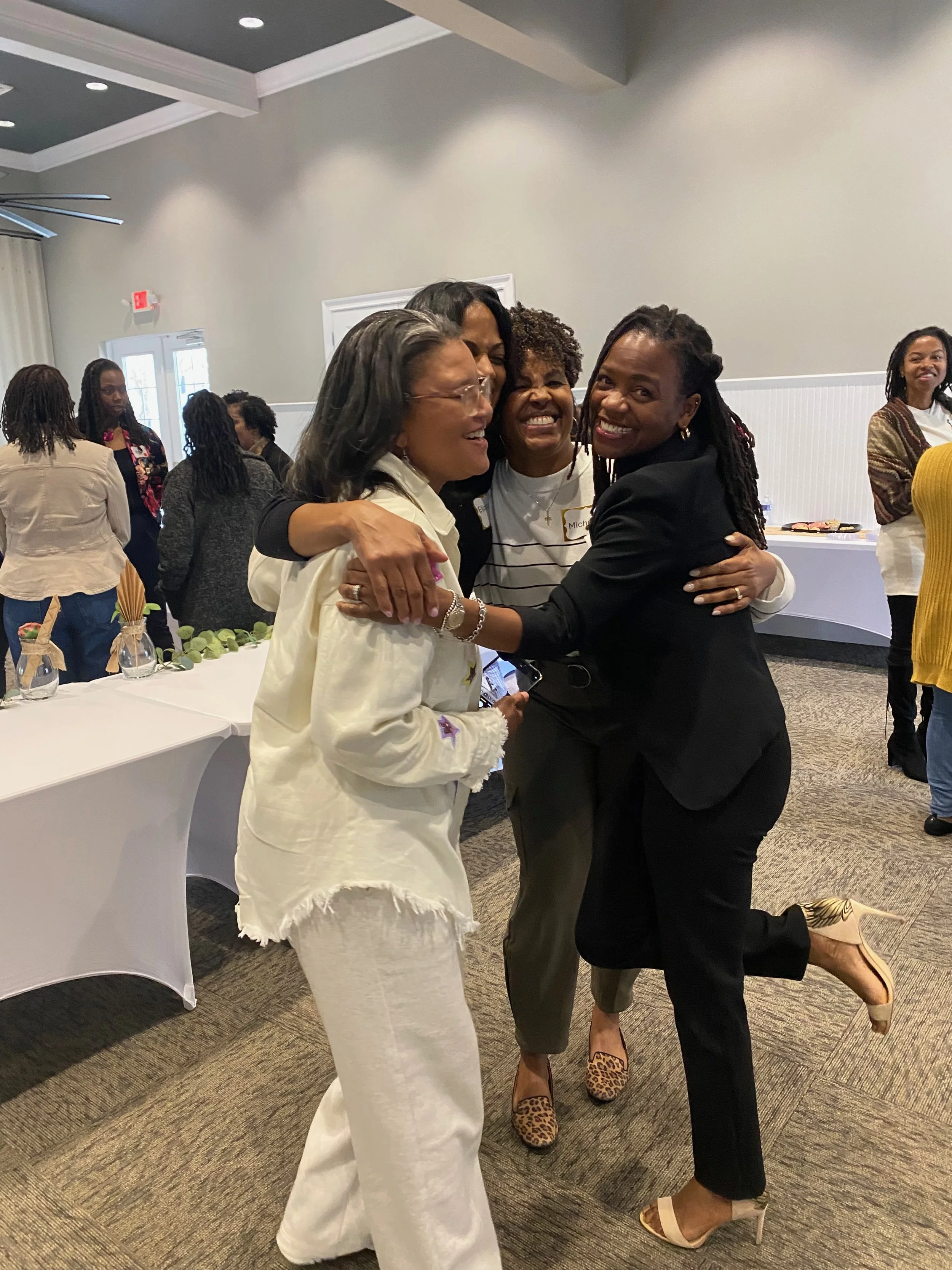Three women are hugging and smiling, with other people in the background at an indoor event. The woman in the middle has curly hair, while the other two women have straight hair, and one is wearing a black blazer with leopard print shoes.