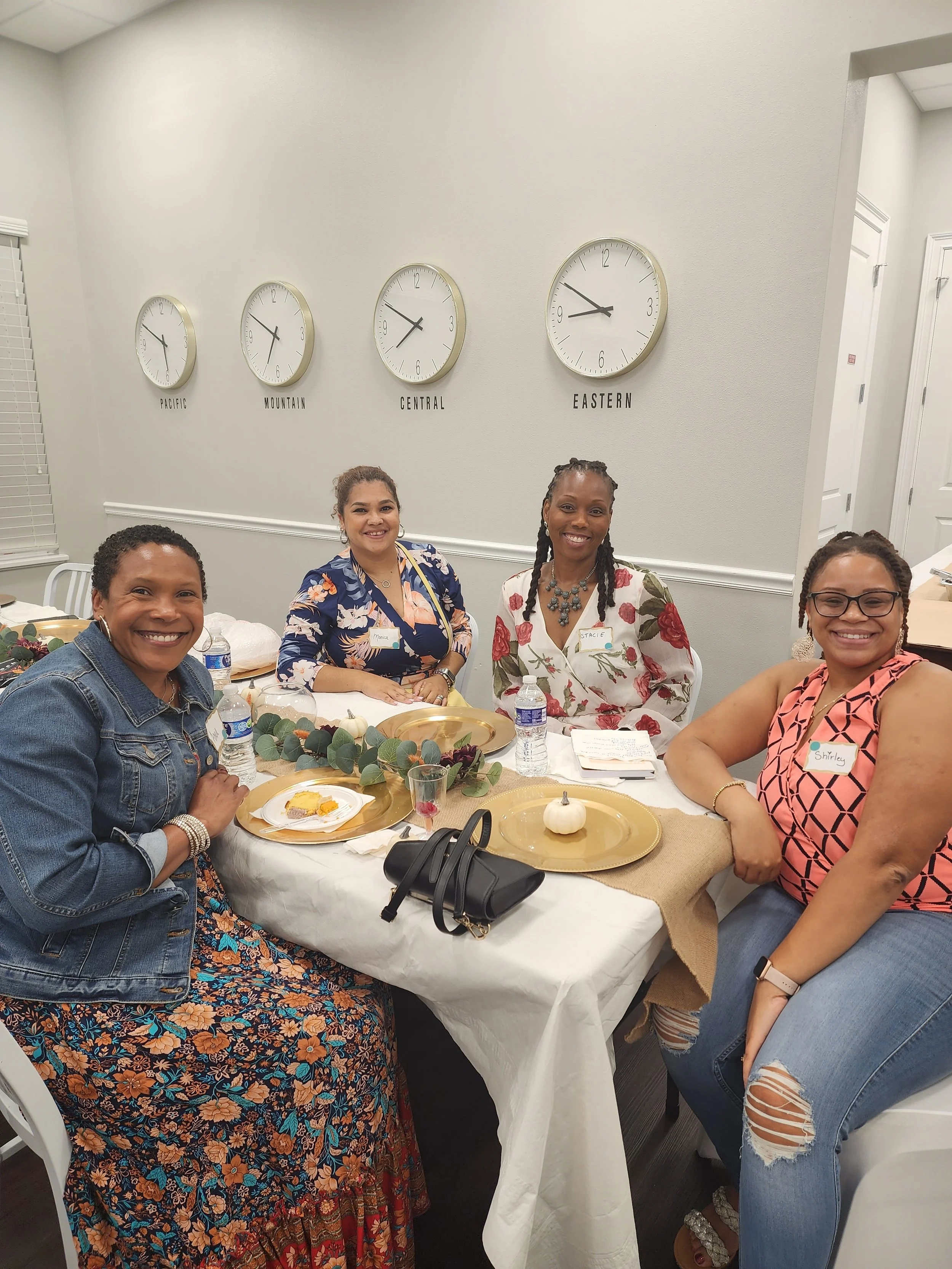 Group of women sitting at a dinner table with gold-rimmed plates, bottles of water, and fall decorations, smiling for the camera, in an indoor setting with clocks showing different time zones on the wall.