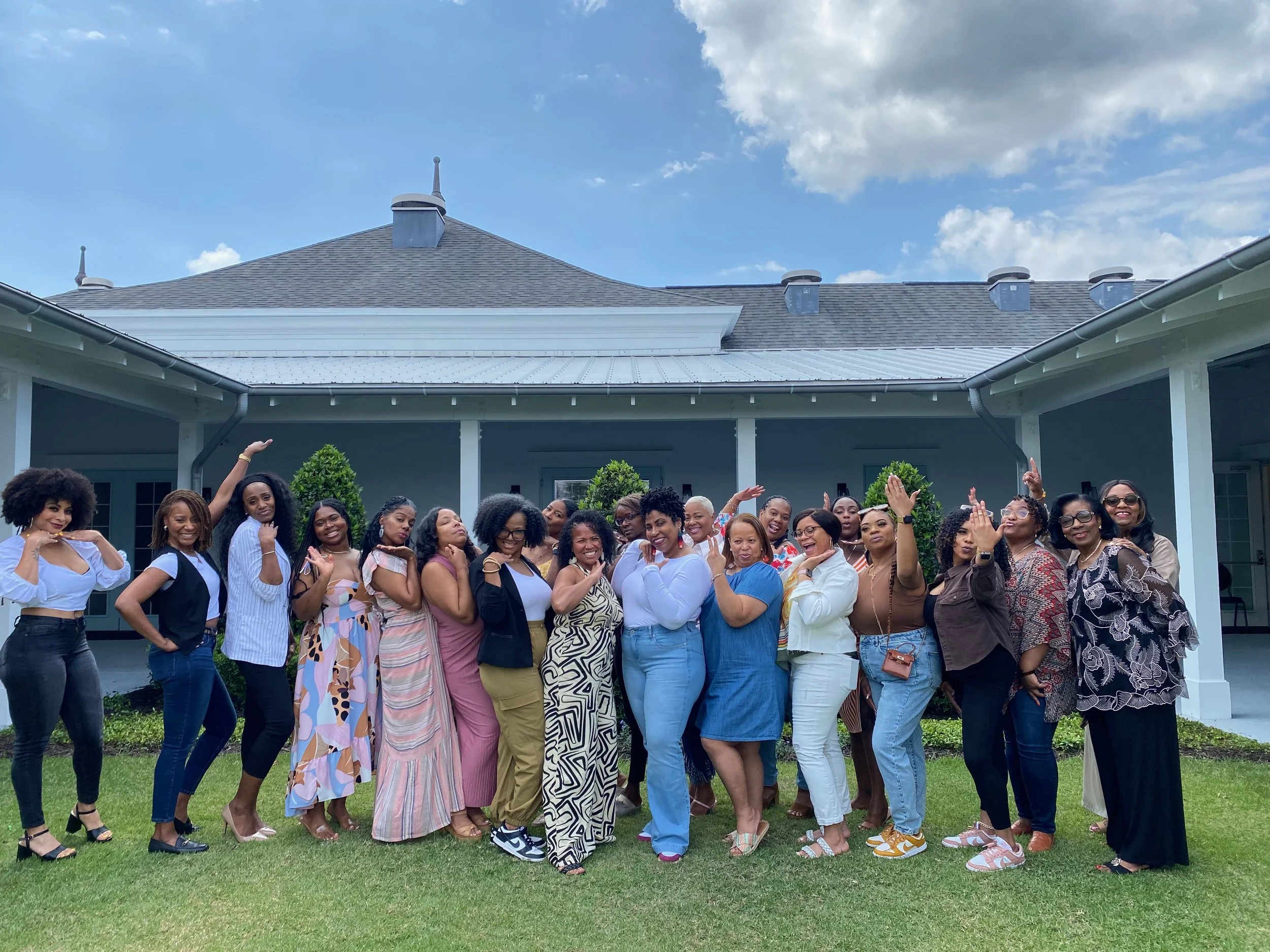 A group of diverse women standing and posing outdoors in front of a house with a gray roof, under a partly cloudy sky. Some women are smiling, and a few have their hands raised or are making gestures.