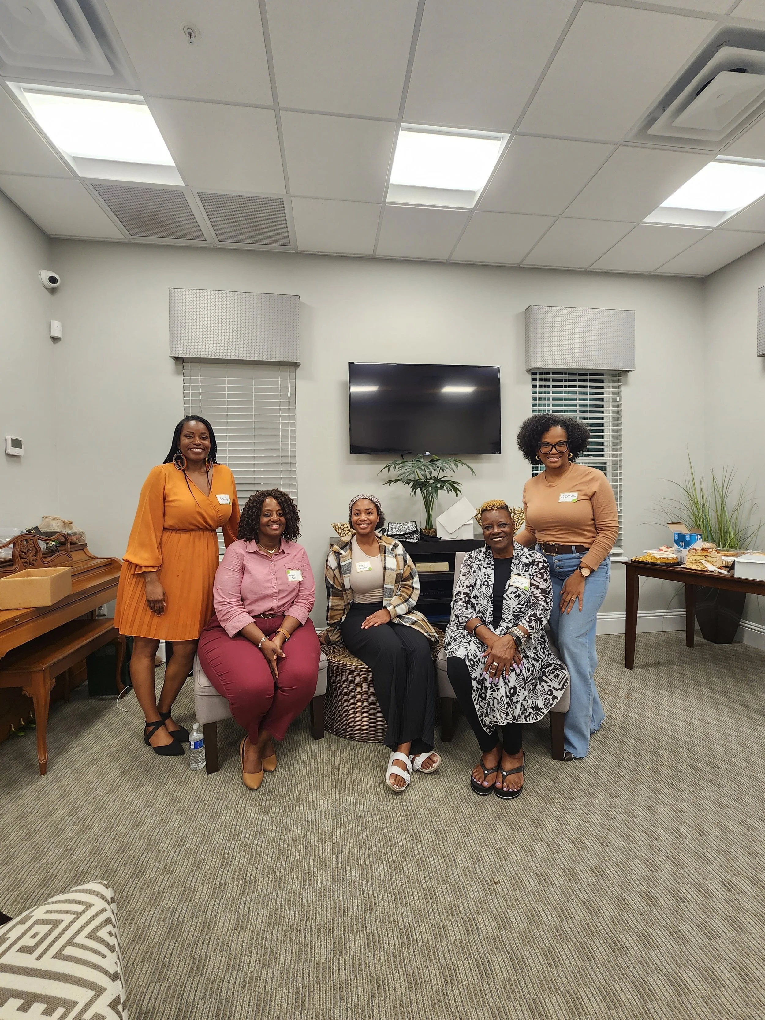 Group of six women gathered in a room, smiling and posing for the photo, with refreshments and a large TV screen in the background.