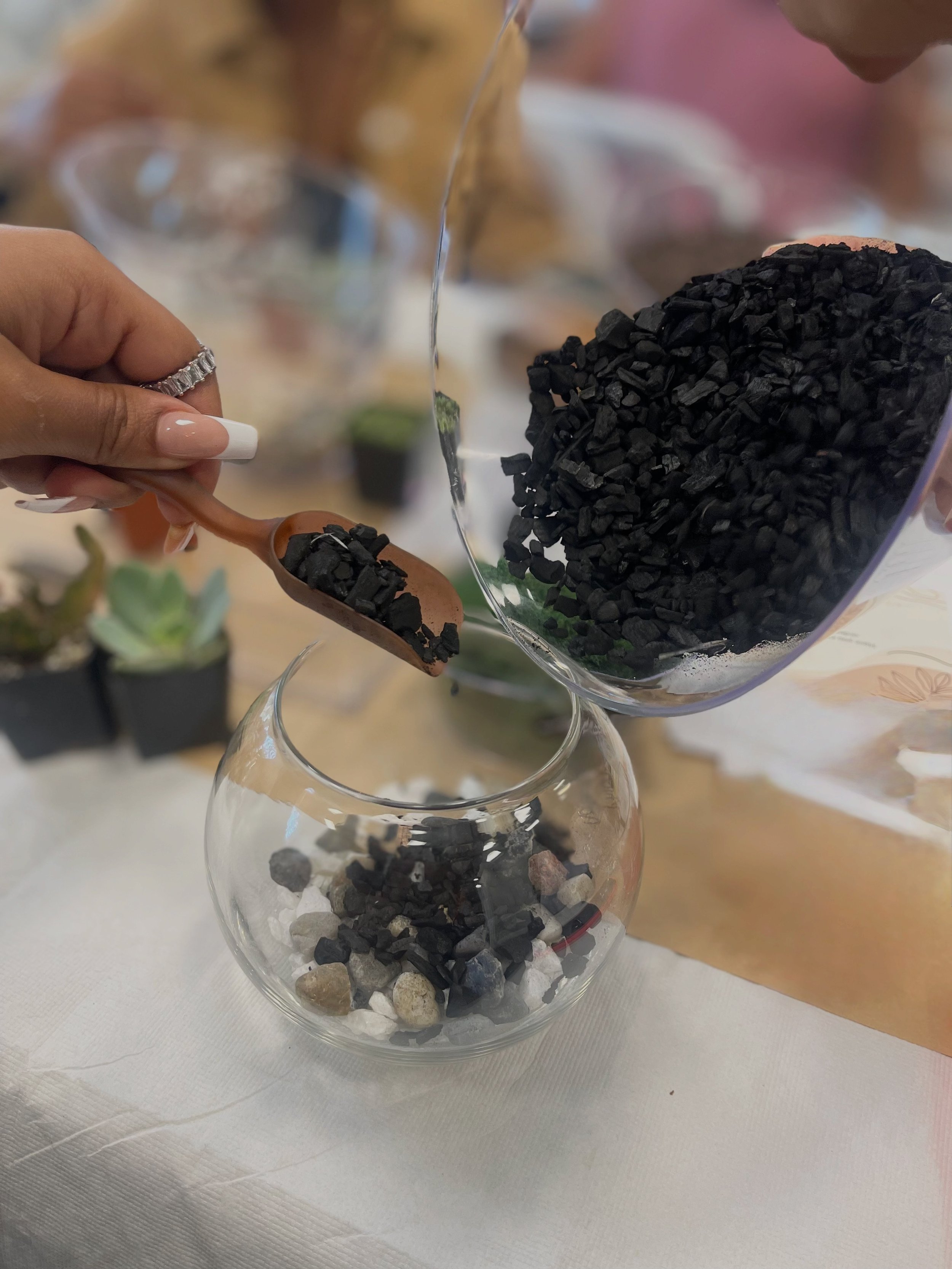 Hand holding a small spoon with black gravel, pouring larger black gravel from a container into a glass vase with a layer of rocks at the bottom, on a white tablecloth surface.