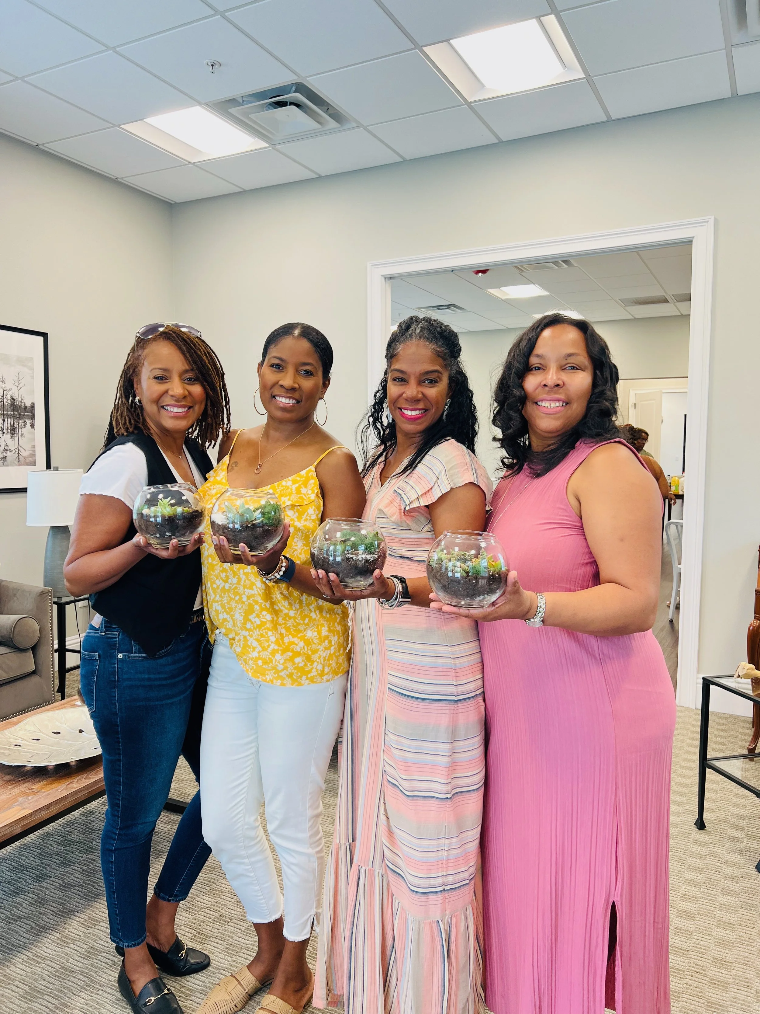 Four women standing together indoors, holding glass bowls with plants, smiling at the camera, with a beige wall, framed picture, and furniture in the background.