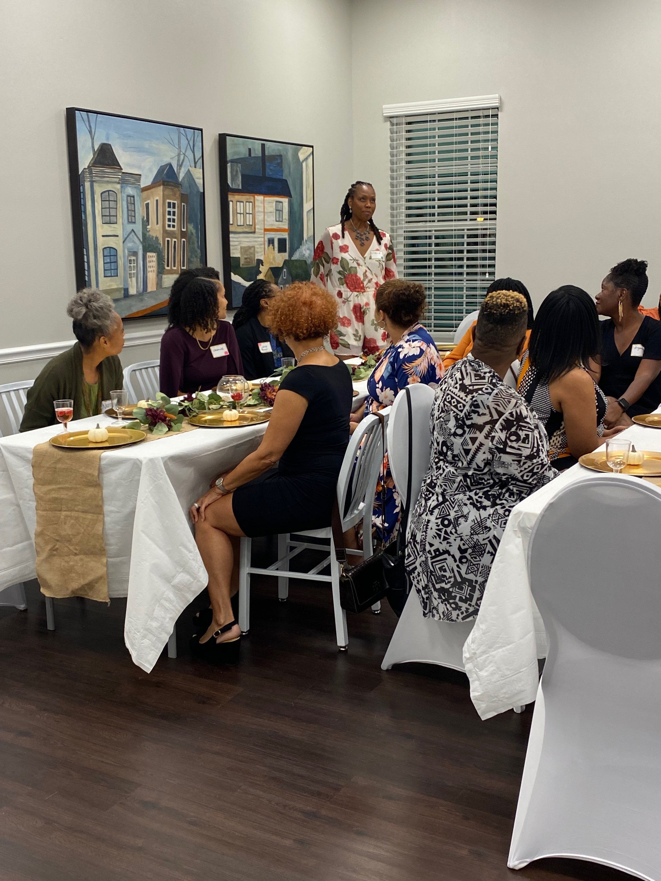 A group of women sitting at a long table with white tablecloths, plates, glasses, and decorations, listening to a woman in a floral dress speaking in a room with gray walls and framed artwork of houses on the wall.