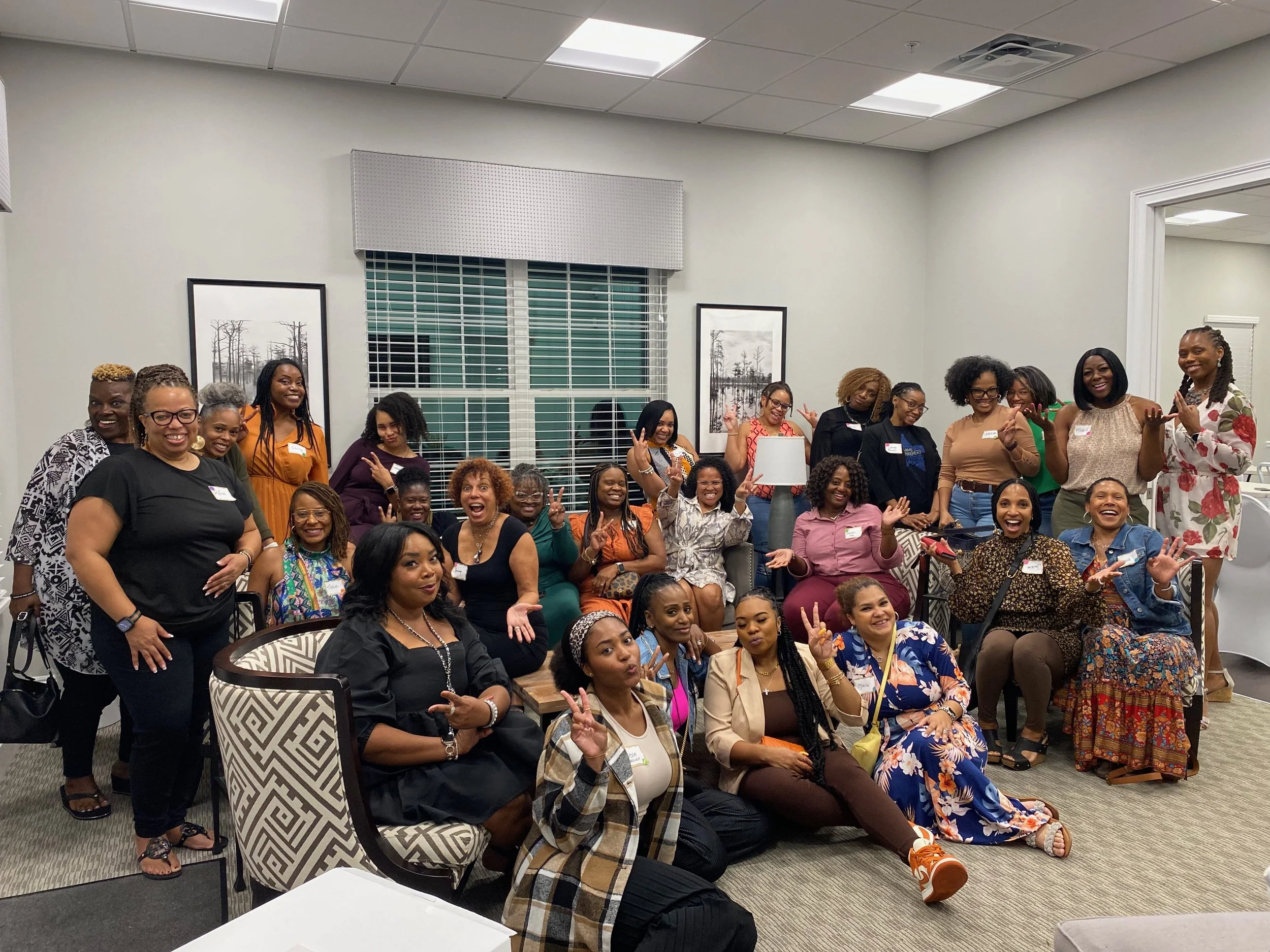 A group of diverse women and girls gathered in a room for a social event or meeting, smiling and posing for the photo. Some are seated, others standing, with a few making peace signs. The room has framed pictures, a window with blinds, and a modern c