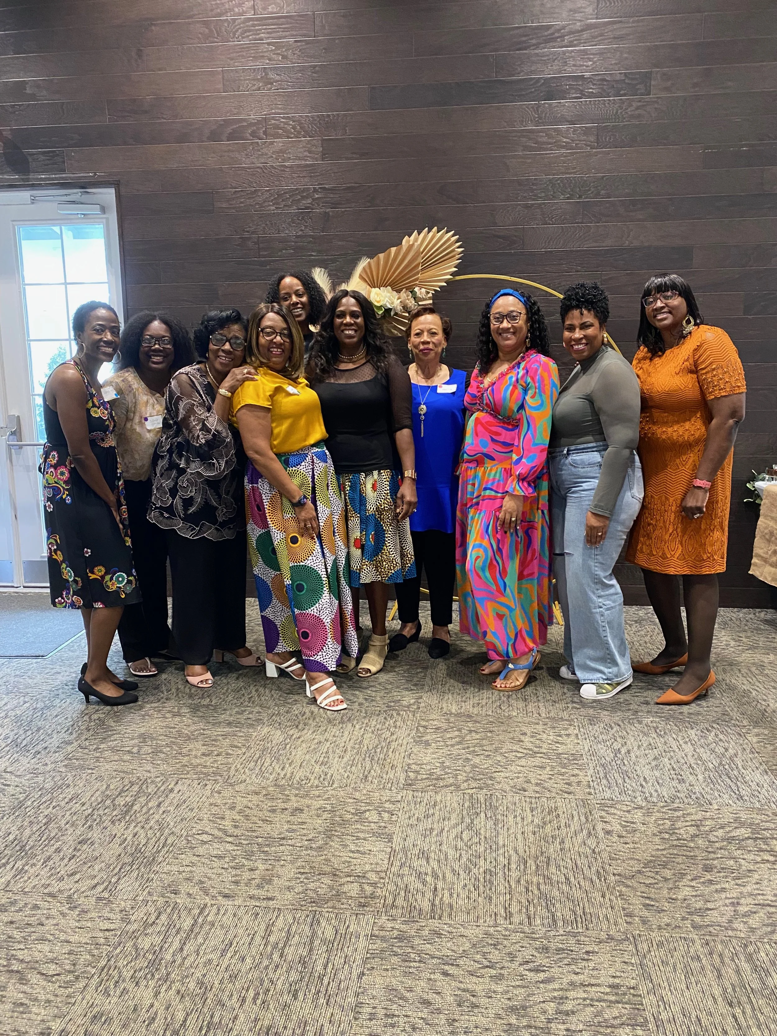 Group of ten women standing together indoors, smiling for a photo, dressed in colorful and stylish clothing, with a decorative floral and fan arrangement in the background on a wooden wall.