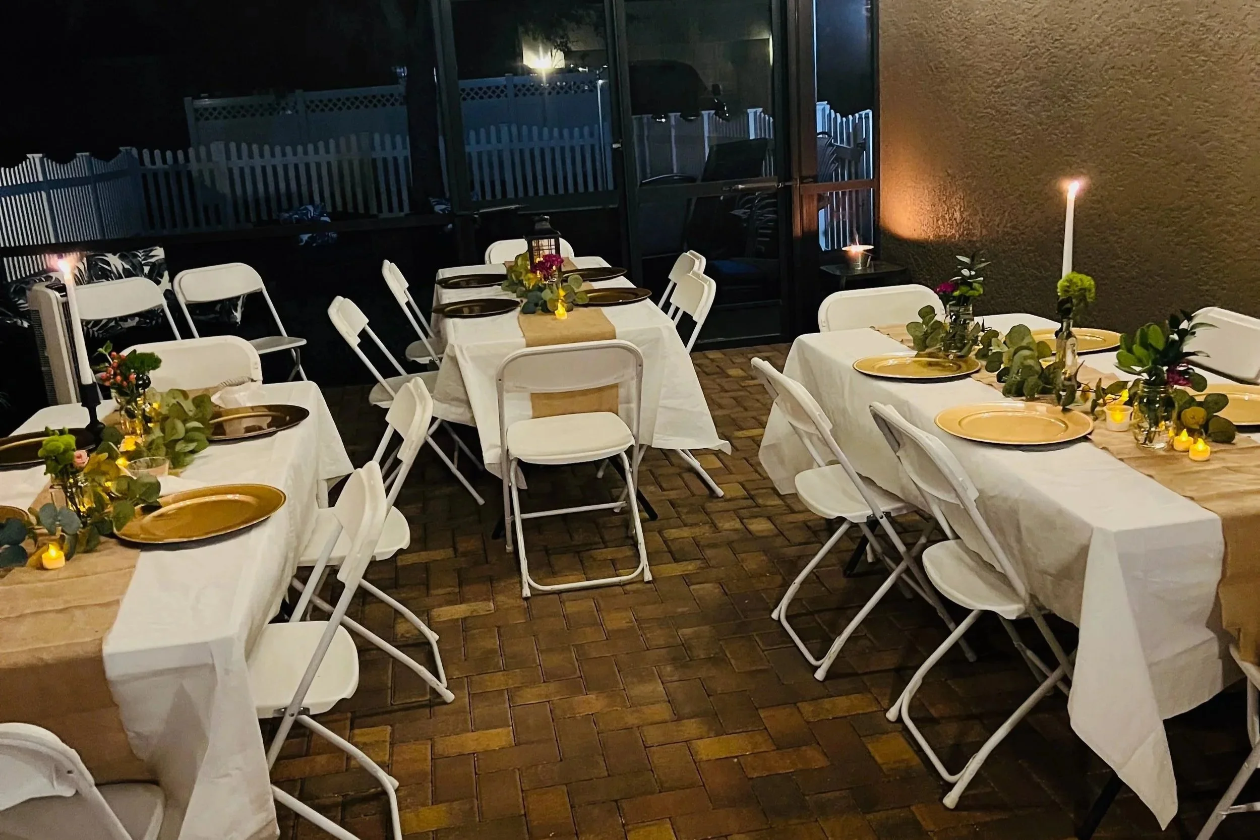 Dinner tables set with white tablecloths, gold plates, green floral centerpieces, and candles, outside at night with a screened porch and fence in the background.