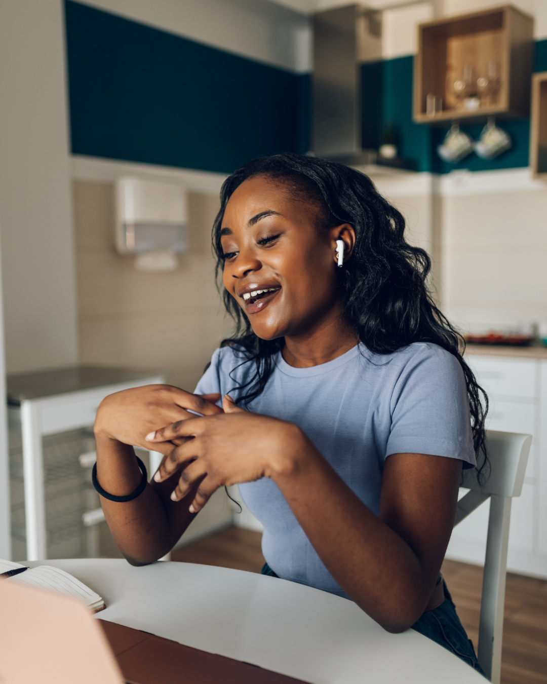 A young woman with black curly hair smiling and talking, wearing a light blue t-shirt and wireless earbuds, sitting at a table in a modern kitchen.