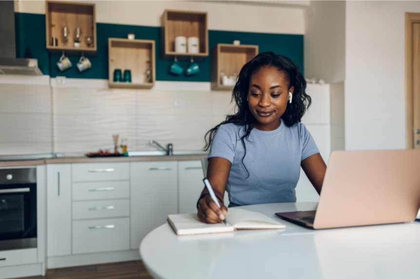 A young woman sitting at a kitchen table writing in a notebook with a laptop open in front of her and earphones in her ears.
