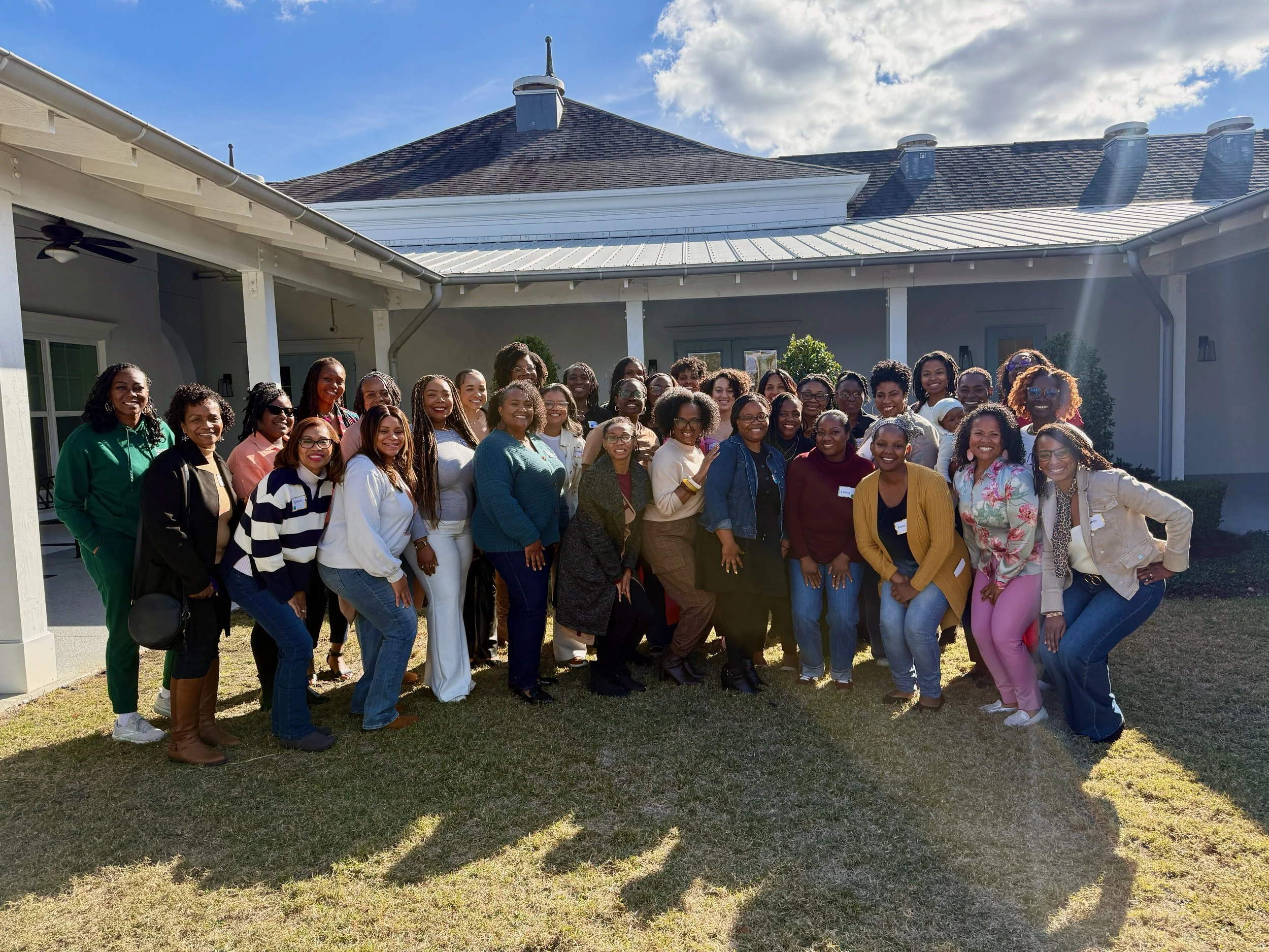 A large group of women gathered outdoors, smiling, in front of a house with a sloped roof and a partly cloudy sky.