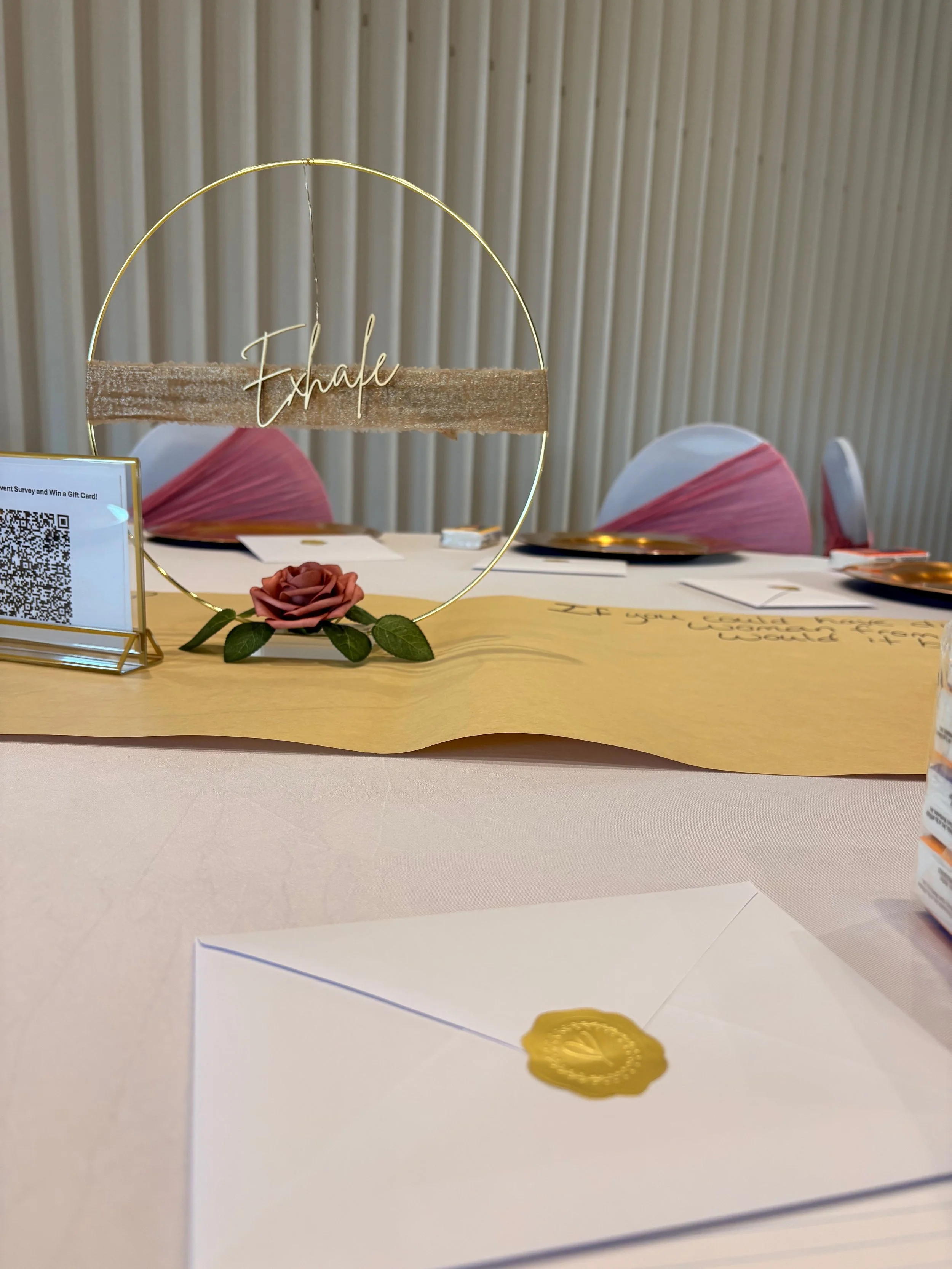 A wedding or formal event table setup with a decorative paper circle, a pink cloth rose, a gold wax seal, and an envelope in the foreground. In the background, there are two chairs with pink and white chair covers and a table with place settings.