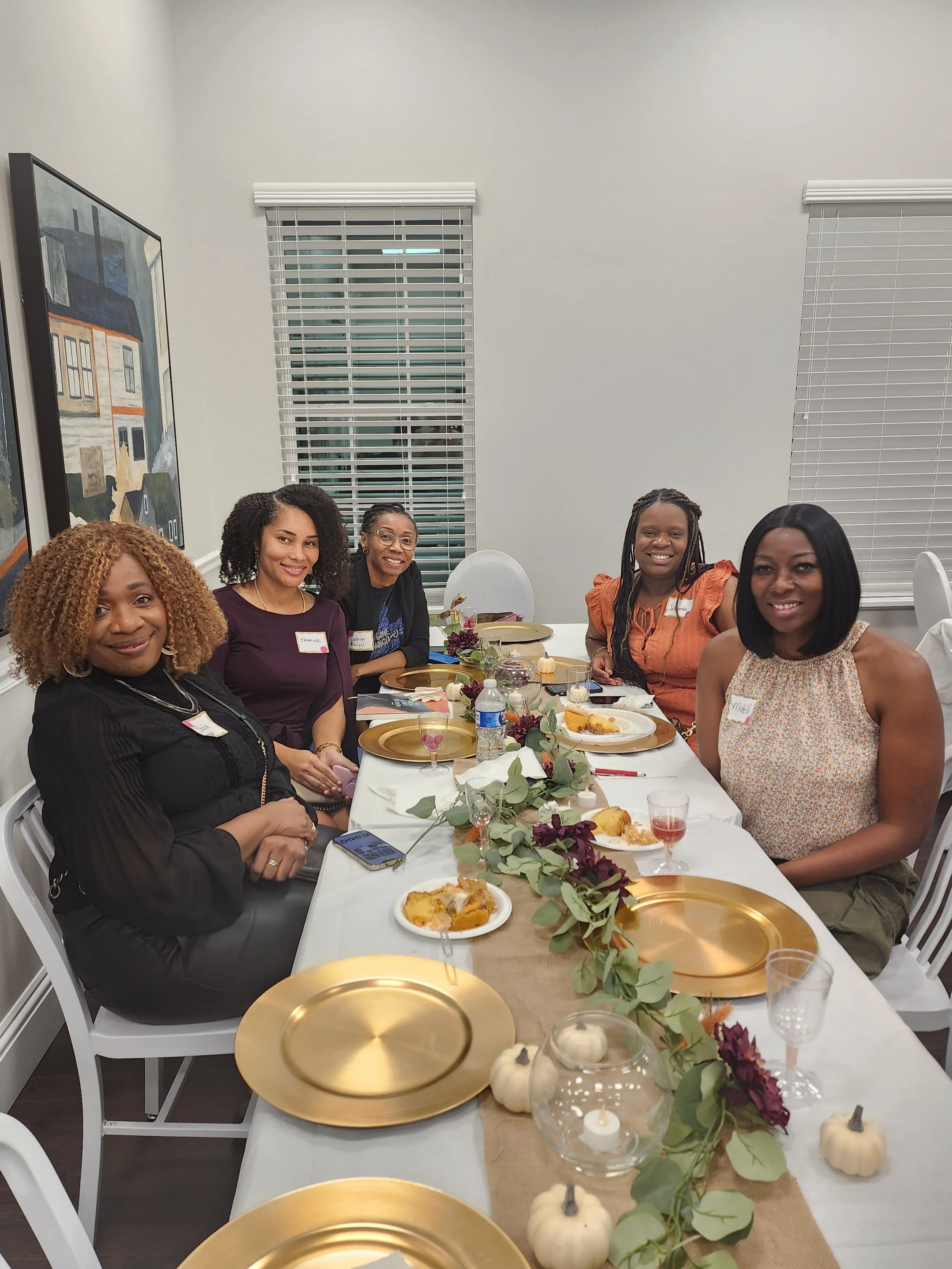 Five women sitting at a decorated table during a meal, smiling at the camera, with plates of food, drinks, and fall-themed decorations including pumpkins and leaves.