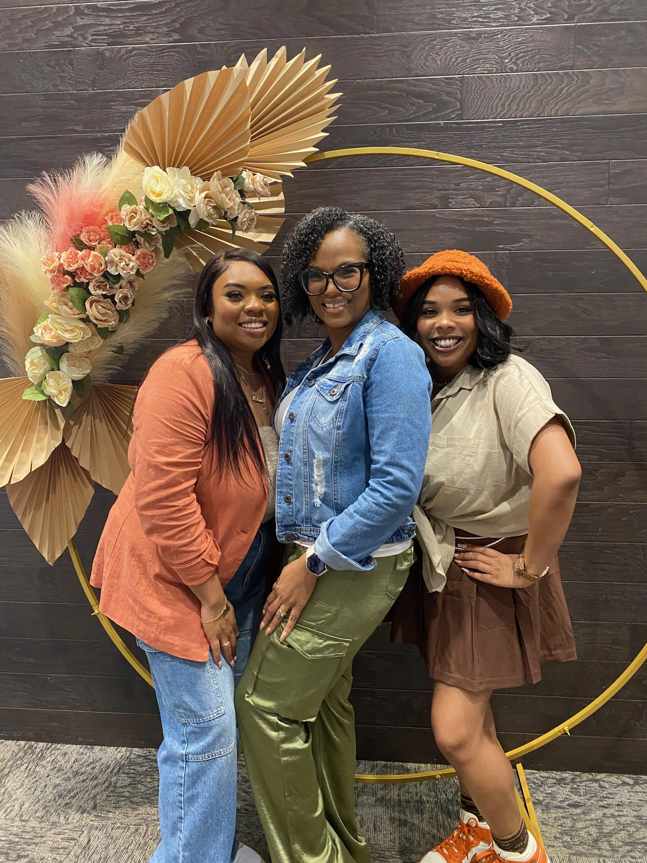 Three women standing together in front of a dark wood wall with a decorative floral and leaf display, all smiling at the camera.
