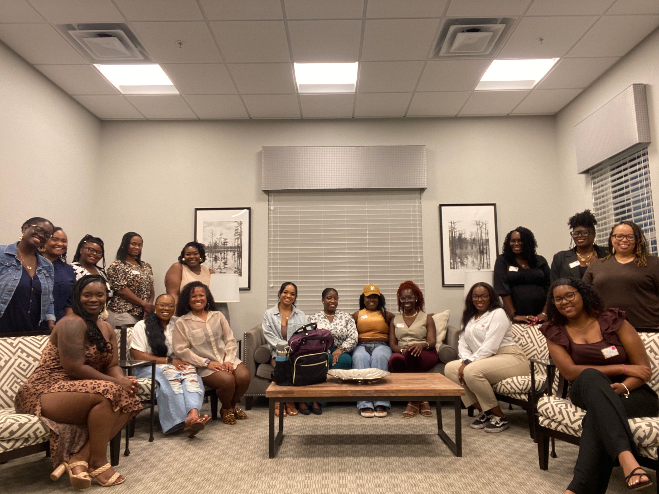 Group of diverse women gathered in a cozy, well-lit room with gray walls, black and white framed photos, and a window with blinds. They are sitting and standing around a table, smiling for the photo.