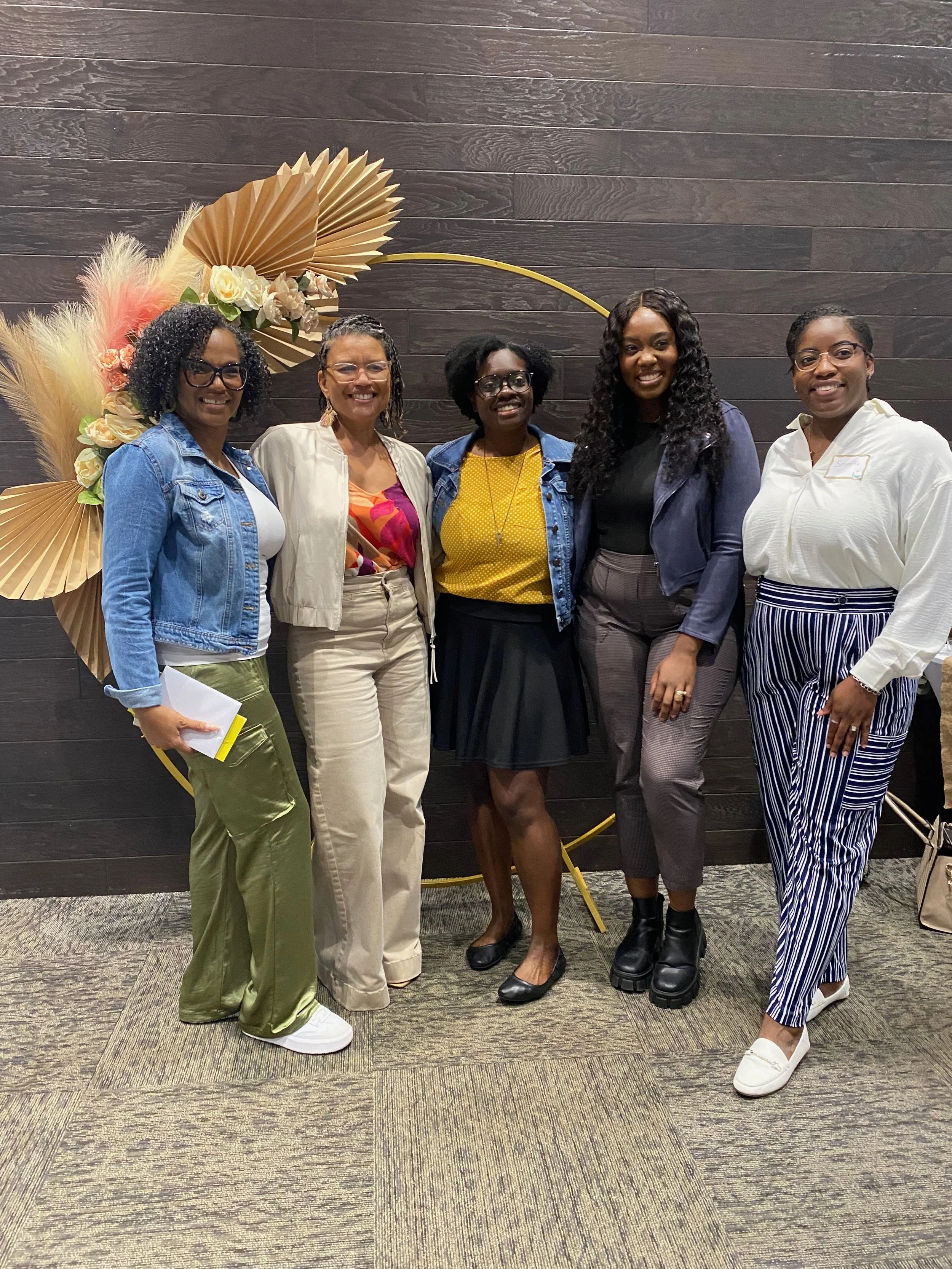 Five women standing together in front of a dark wooden wall with decorative dried palm leaves and flowers. They are smiling and dressed in various casual and business outfits.