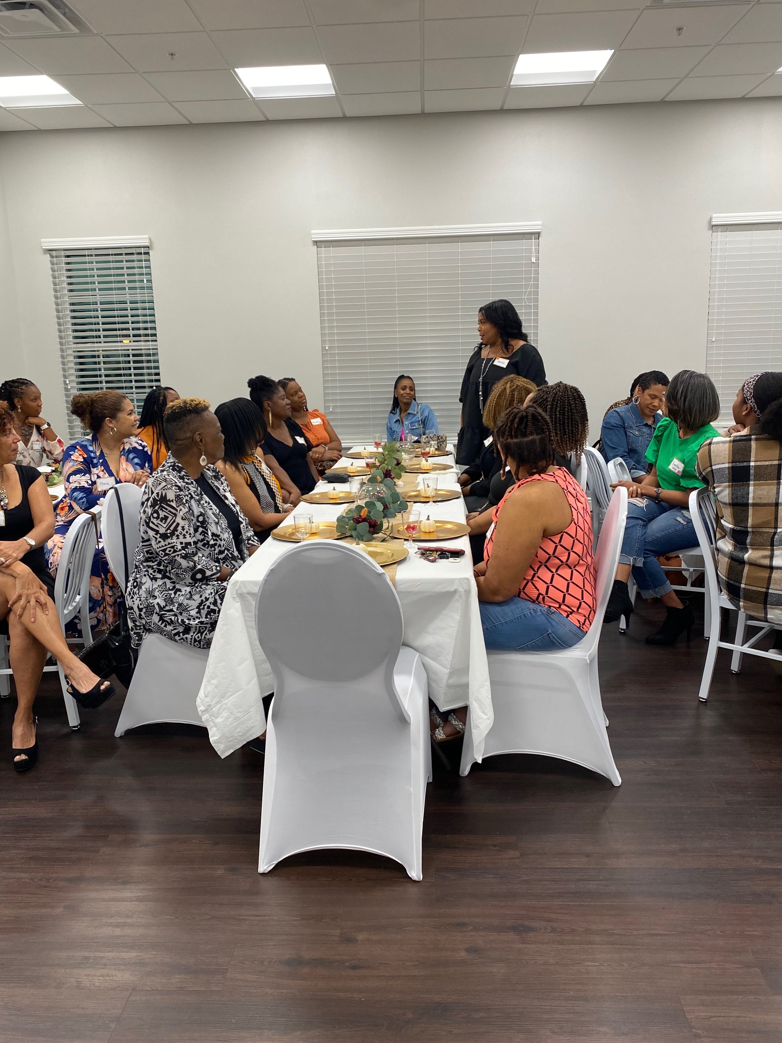 A group of women seated around a banquet table in a well-lit room, with a woman standing and speaking to them.
