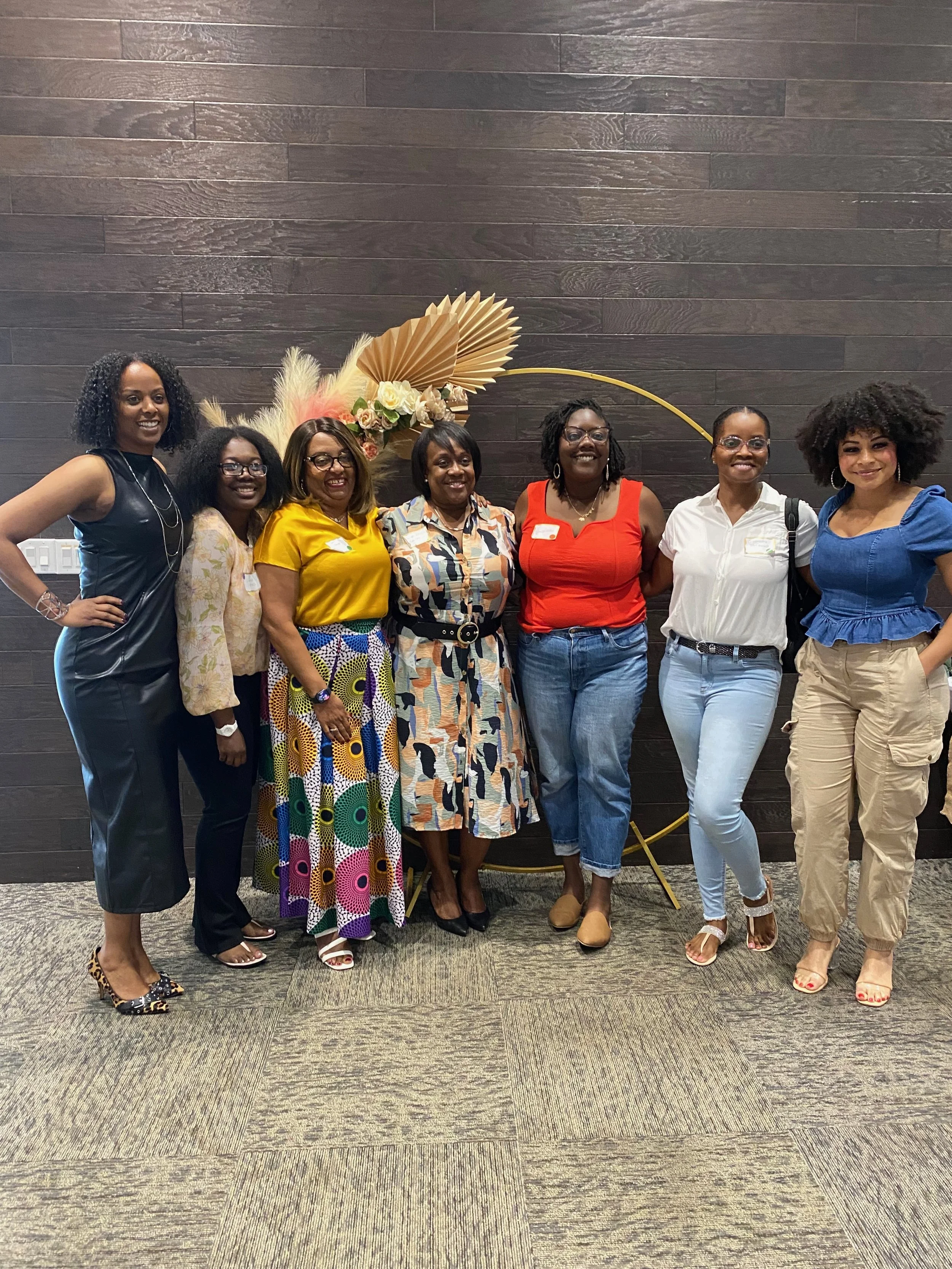 A group of seven women standing side by side in front of a dark wooden wall and a floral decoration, smiling at the camera.