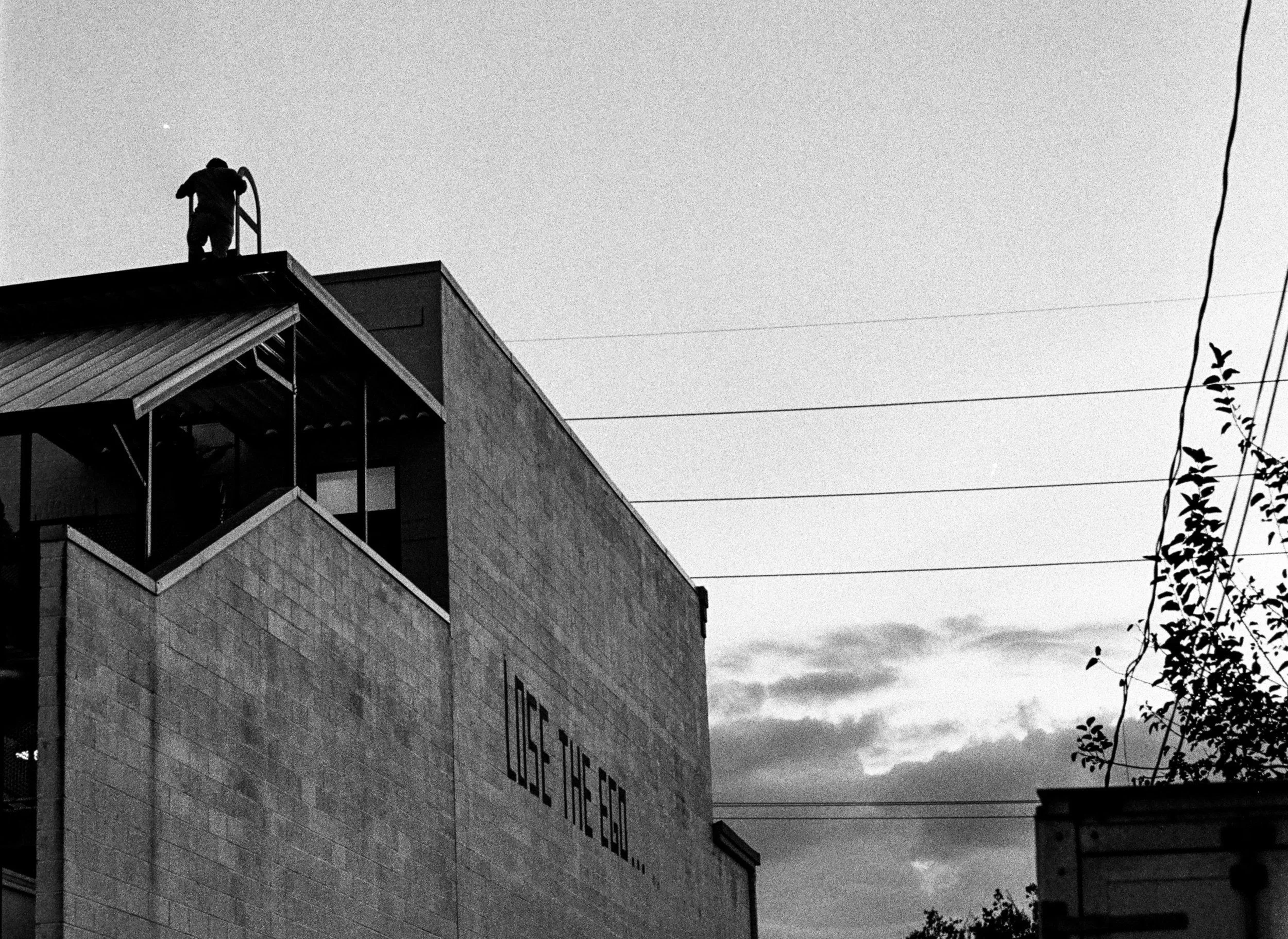 Black and white film photo of a brick building painted with the words Lose the Ego, a figure climbing onto the roof at dusk