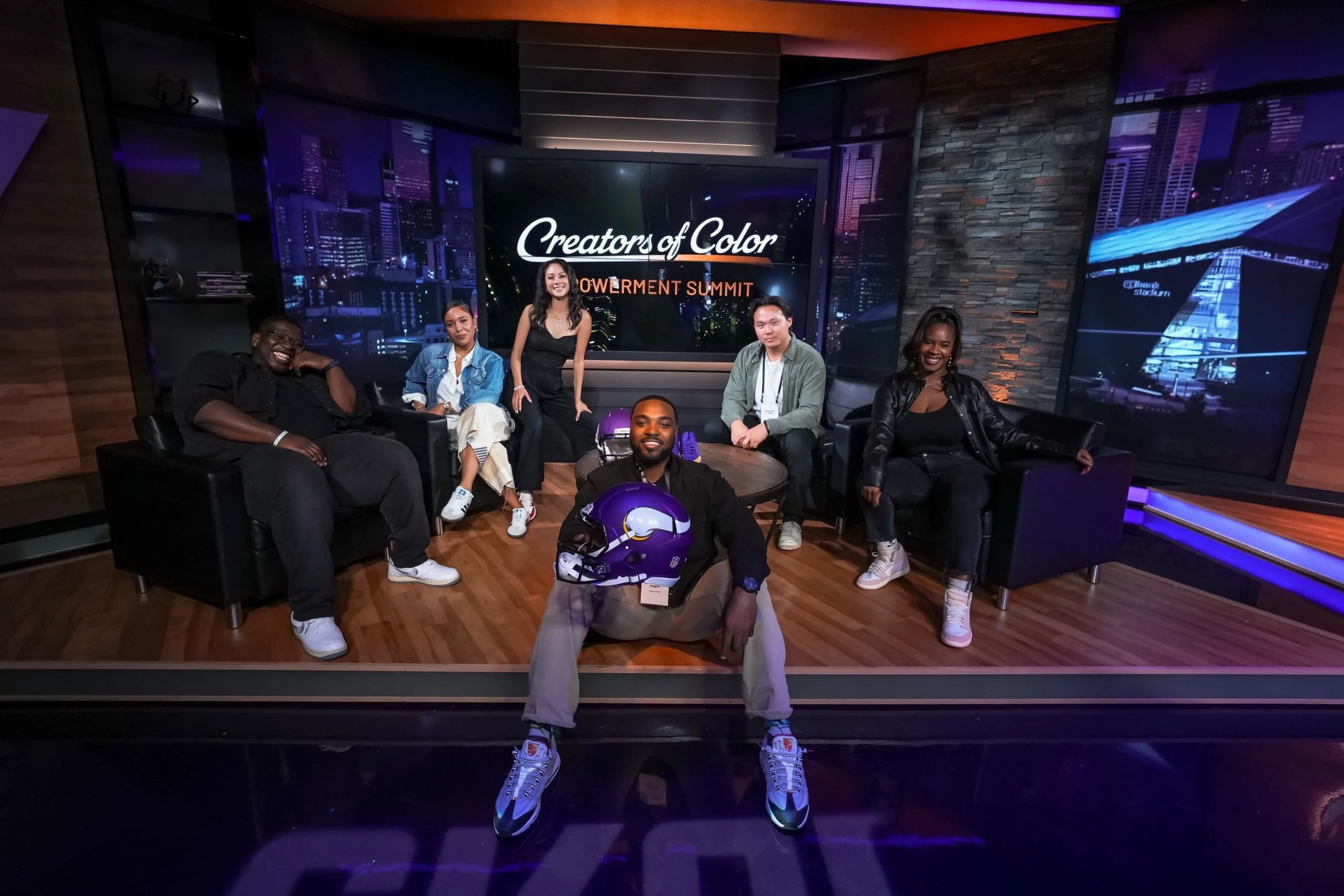Group of six diverse people on a TV set with a cityscape background, sitting and kneeling around a table, with a screen behind them reading 'Creators of Color Powerment Summit.'