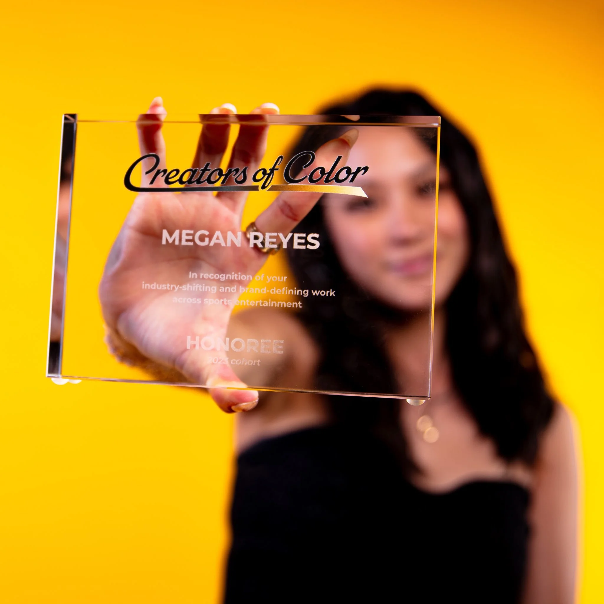 A woman with dark hair holding a clear glass award plaque labeled "Creators of Color" with the name "Megan Reyes" in focus, against a yellow background.