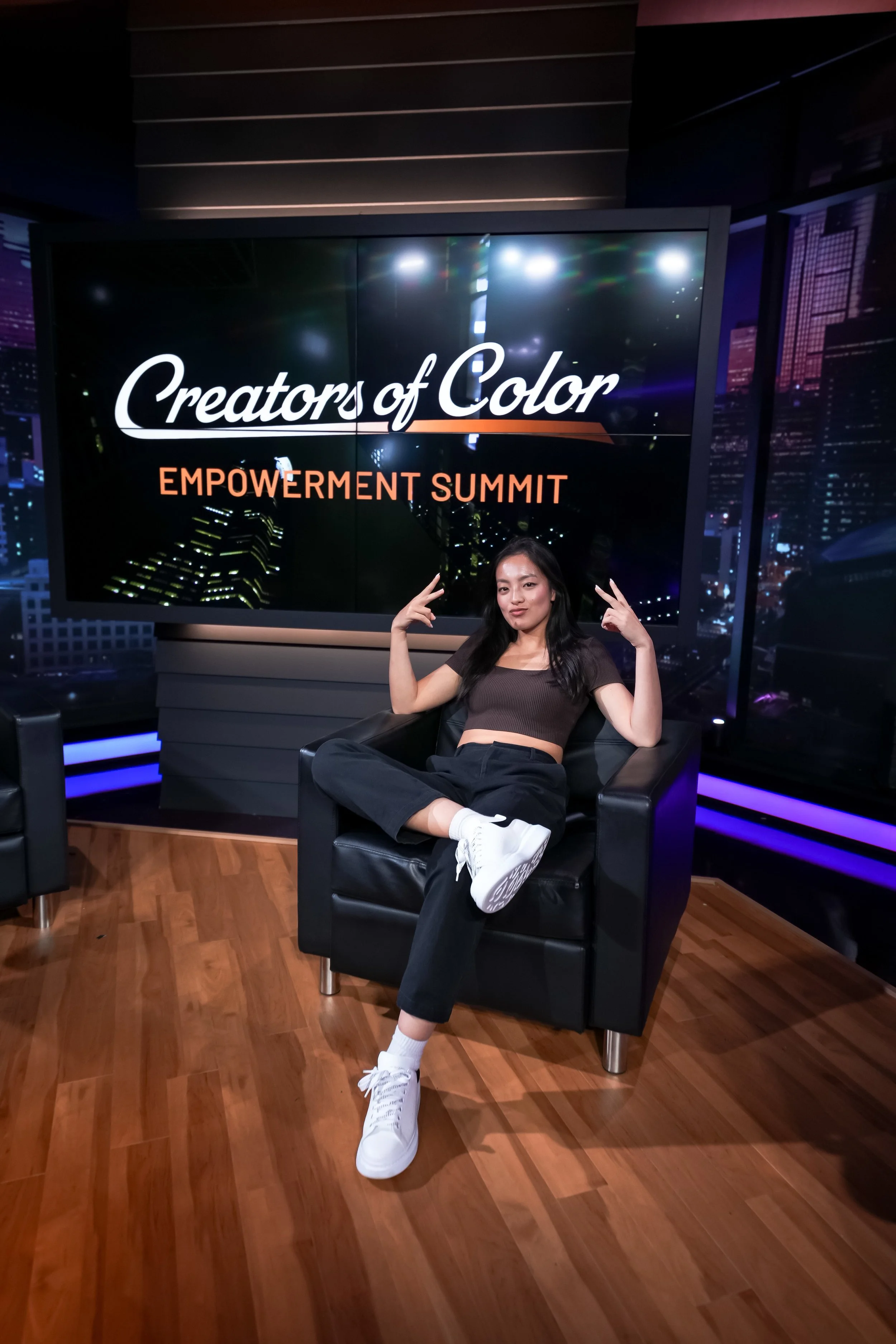 A young woman sitting on a black armchair in front of a large screen that reads 'Creators of Color Empowerment Summit.' She is posing with both hands making peace signs and has a relaxed expression.