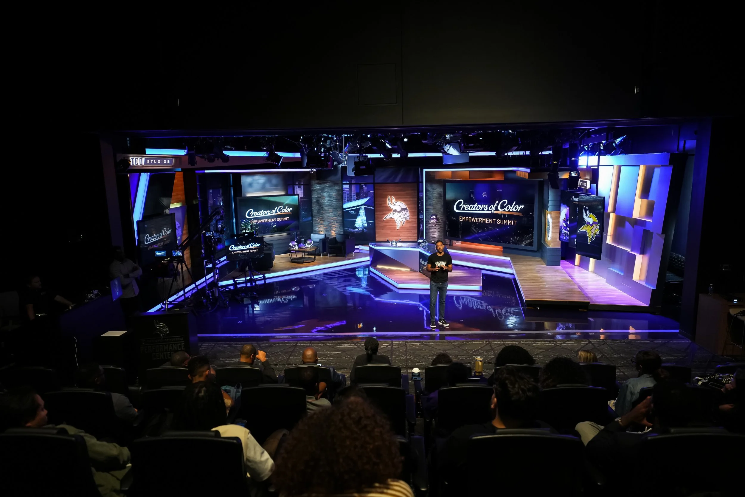 Stage set for the Creators of Color Empowerment Summit with a Minneapolis Vikings logo and Vikings mascot, audience seated facing the stage, stage lit with blue and purple lighting, presenter standing in front of screens.