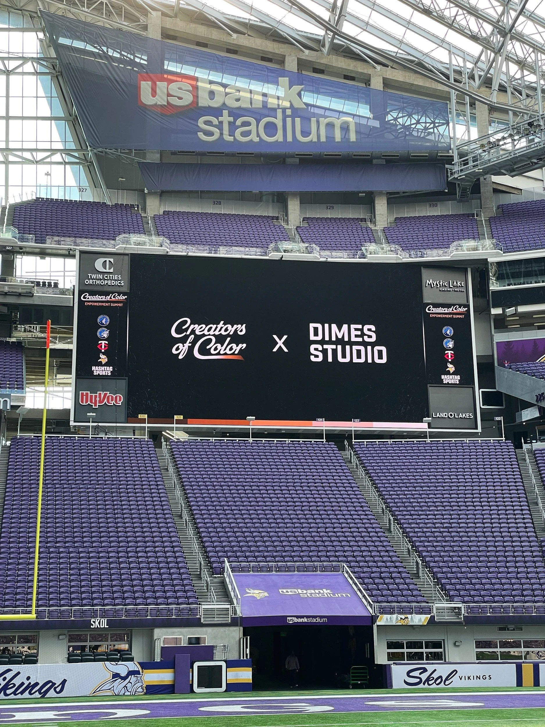Interior view of U.S. Bank Stadium showing a large digital screen with the text 'Creators of Color x Dimes Studio'. The stadium seats are purple, and the field area is visible in the foreground with purple and green turf.