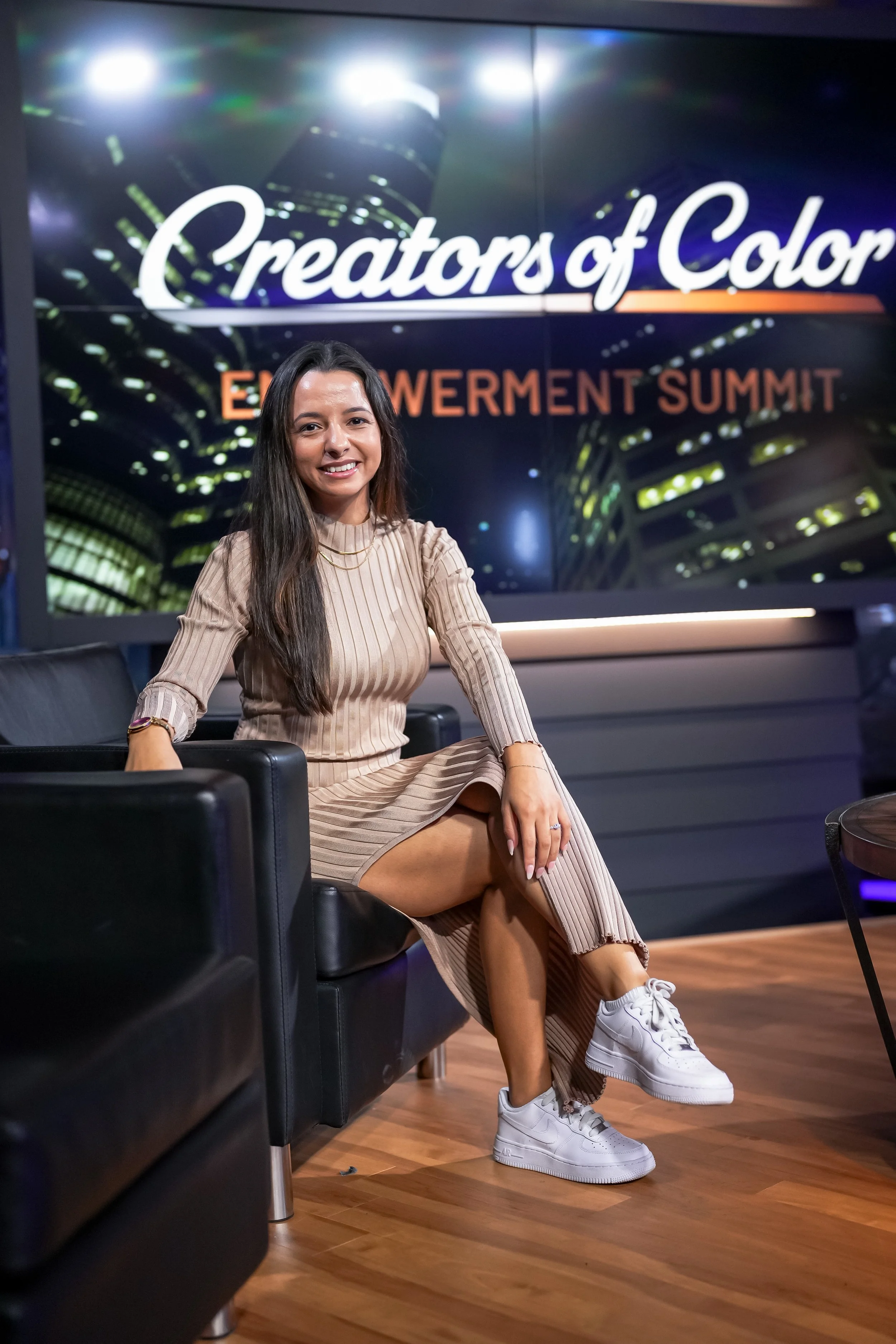 A woman sitting on a black leather chair, smiling, at a professional event with a large screen behind her displaying the words 'Creators of Color' and 'Empowerment Summit' in the background.