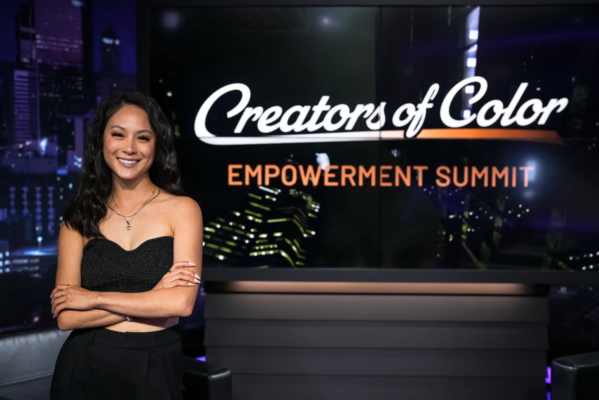 A woman with long black curly hair wearing a black strapless top and black pants, smiling with crossed arms, stands in front of a large screen displaying the text 'Creators of Color EMPOWERMENT SUMMIT' for an event.