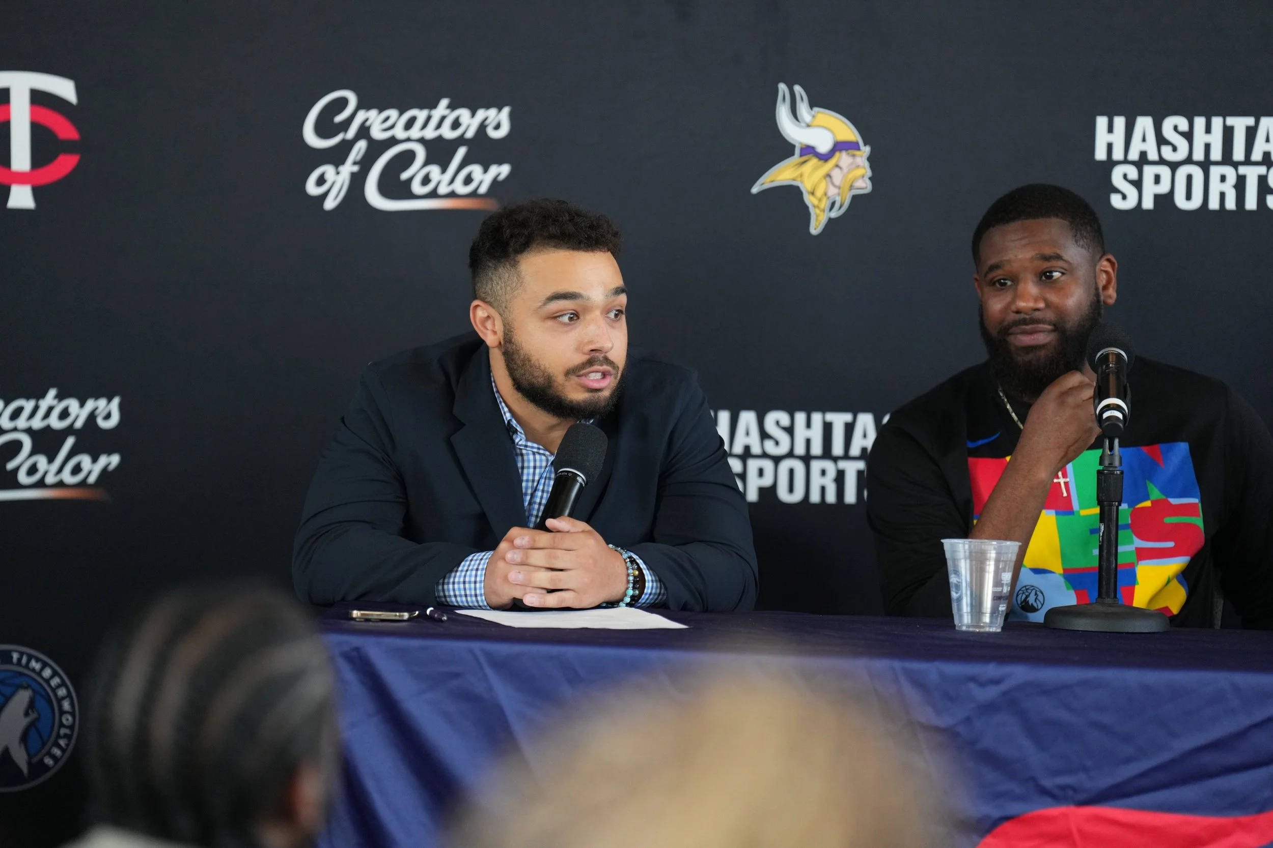 Two men sitting at a table with microphones during a press conference, with a backdrop featuring the Minnesota Vikings logo, the Twins logo, and the phrases "Creators of Color" and "Hashtag Sports."