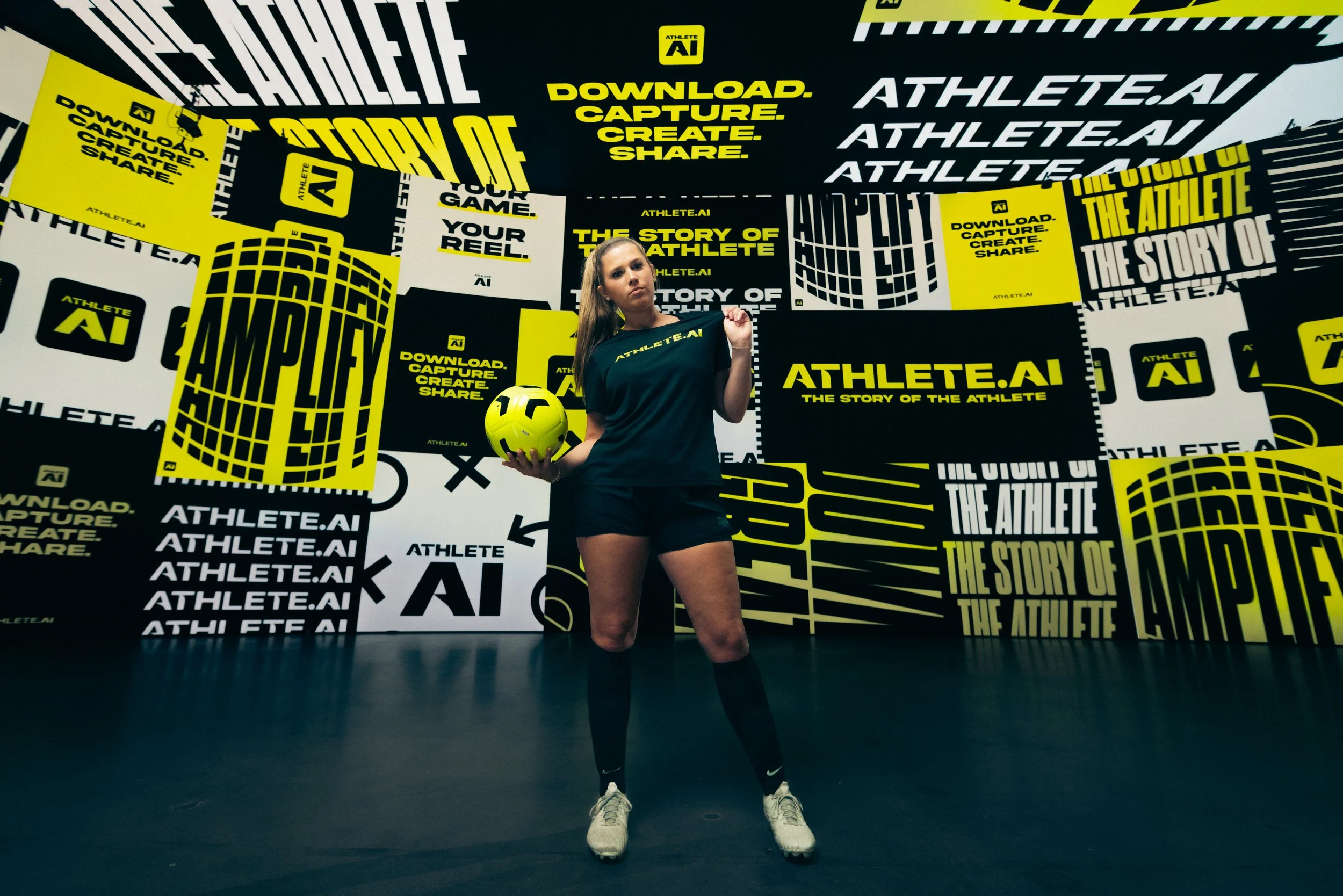 A young woman in athletic clothing standing in a studio with a yellow and black background, holding a yellow soccer ball, wearing a black shirt with 'ATHLETE.AI' printed on it.