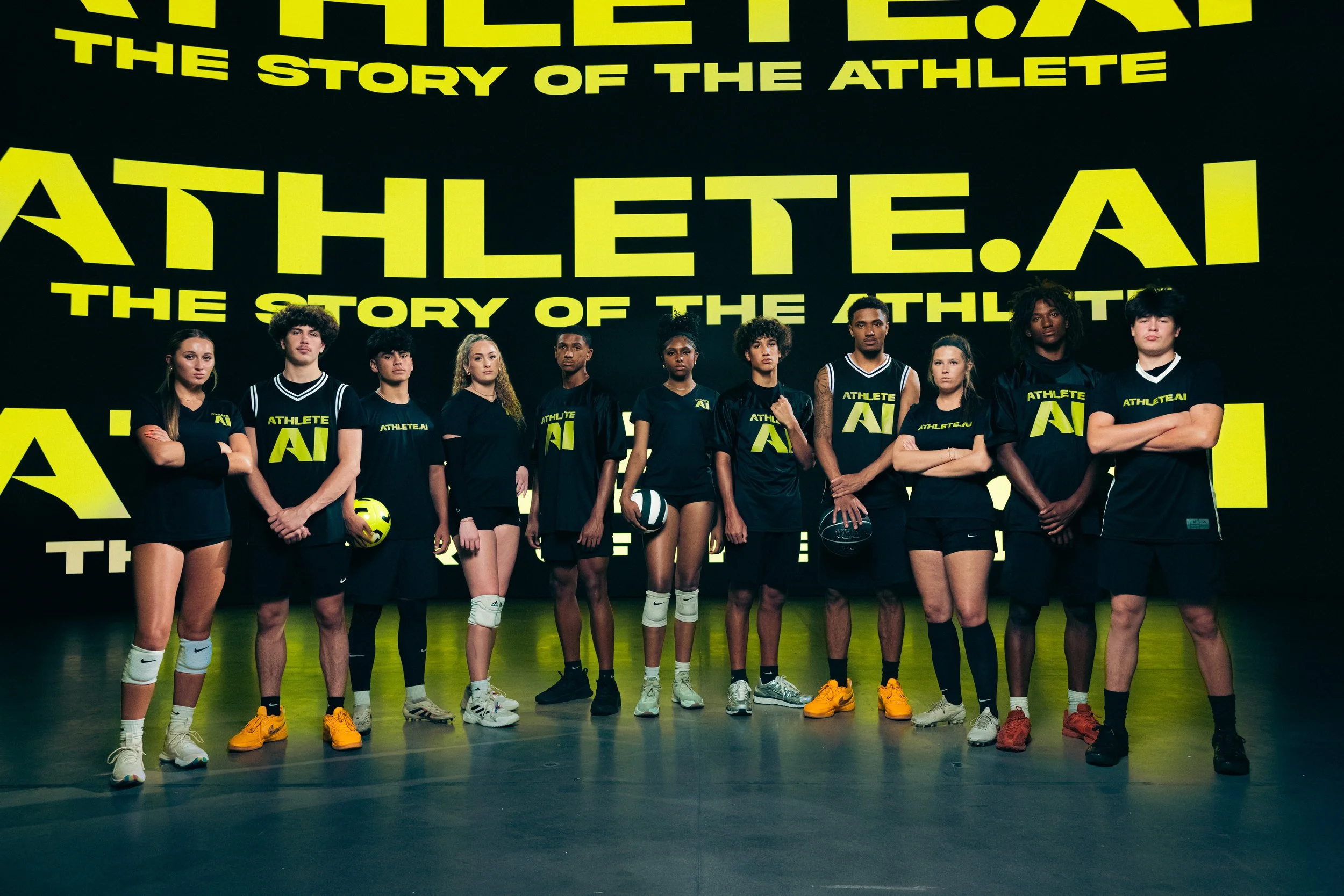 A diverse group of eleven young athletes standing in a line, wearing black athletic clothing with yellow logo and text, against a large black and yellow backdrop with the words 'Athlete.ai' and 'The story of the athlete'.