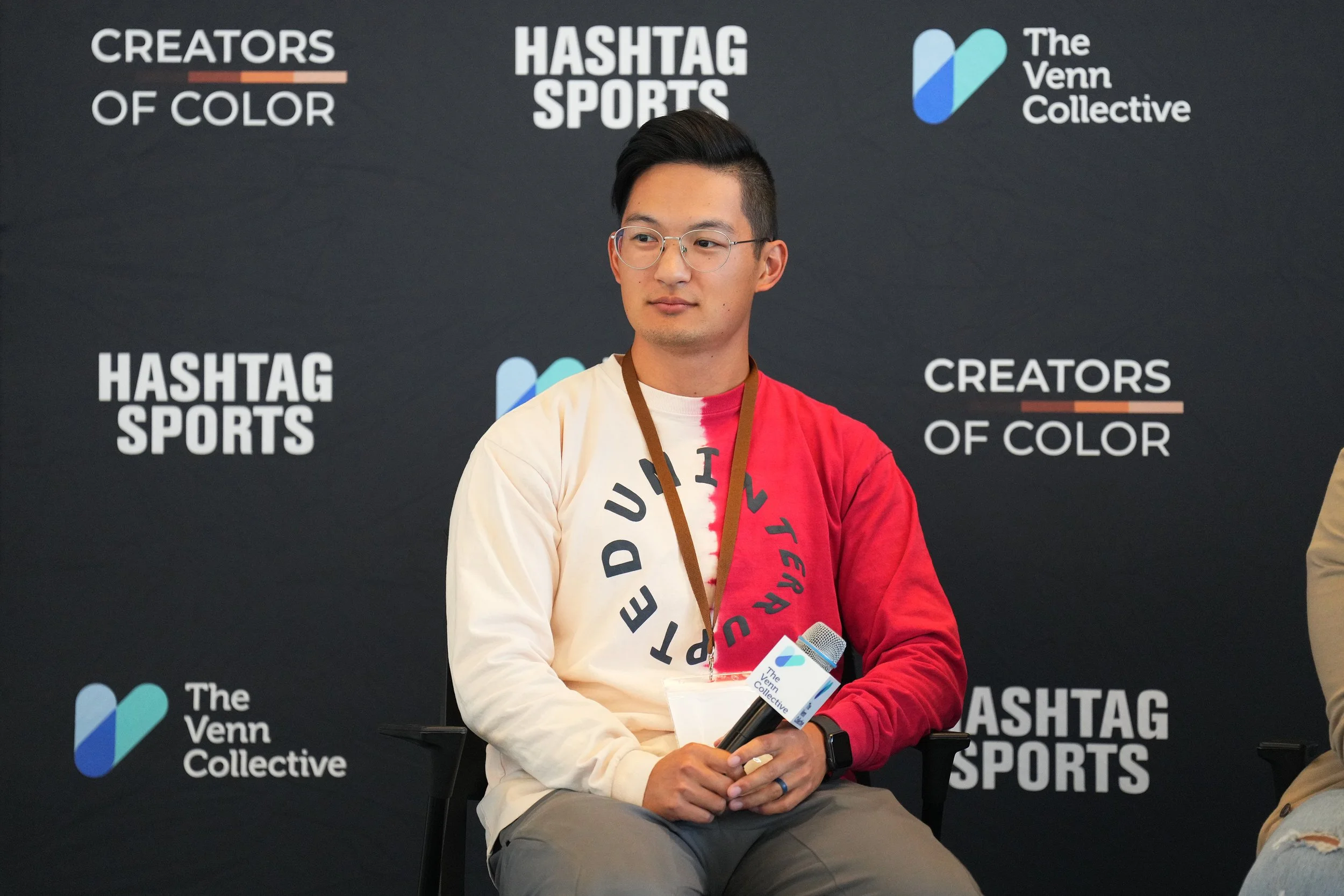 A young man sitting in front of a dark backdrop with logos and text, holding a microphone with a logo, wearing a two-tone red and white sweatshirt with black text, glasses, and a smartwatch.