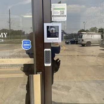 Glass storefront door with QR code notices, security camera sign, keypad access panel, and parking lot reflected in the glass