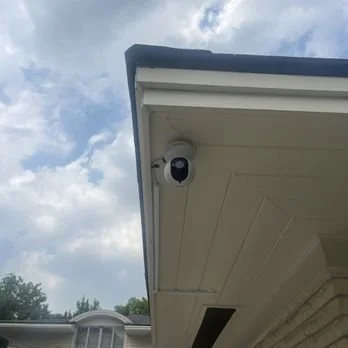 Security camera mounted under a house roof eave against a partly cloudy sky