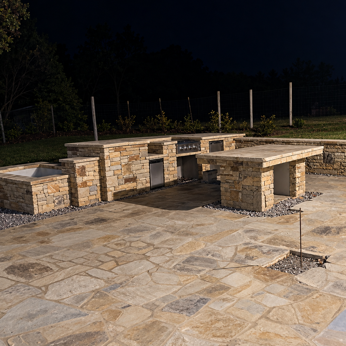 Stone outdoor kitchen with built-in grill, refrigerator, sink, and bar counter on a large flagstone patio at night