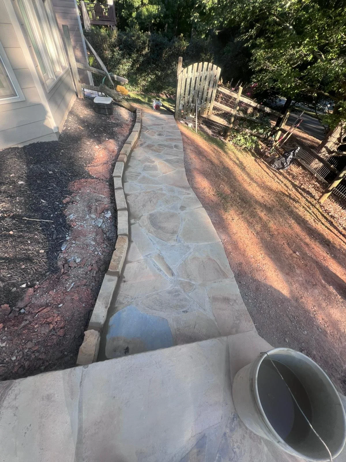 Flagstone walkway with stone edging beside a house, garden beds, wooden gate, and fenced backyard.