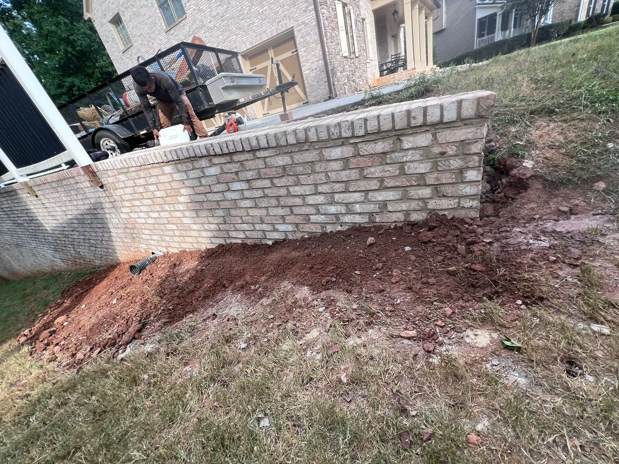 Worker excavating soil beside a residential brick retaining wall, with a utility trailer and construction tools nearby
