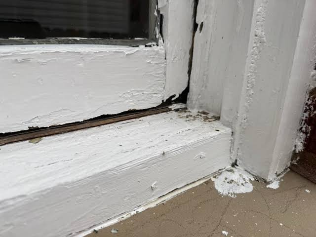 Close-up of peeling white paint and deteriorating wood around a window sill and frame.