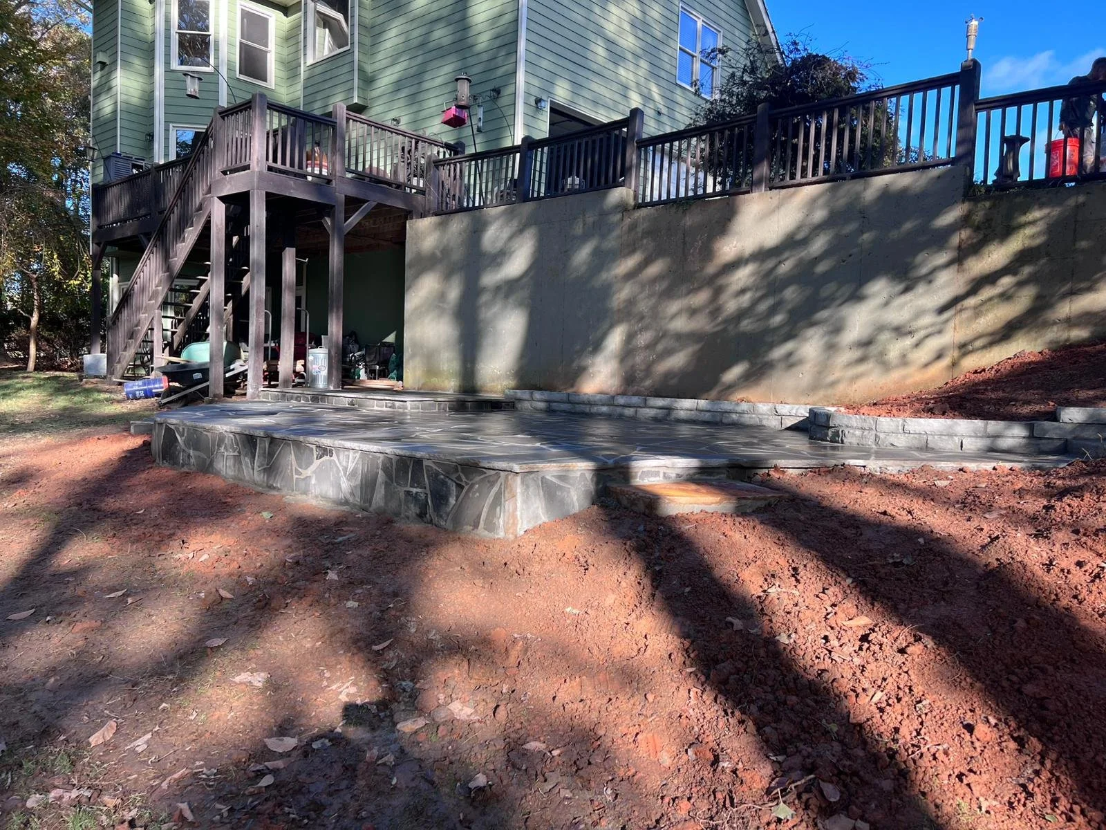 Backyard patio with dark stone paving, retaining wall, wooden deck, stairs, and green house exterior