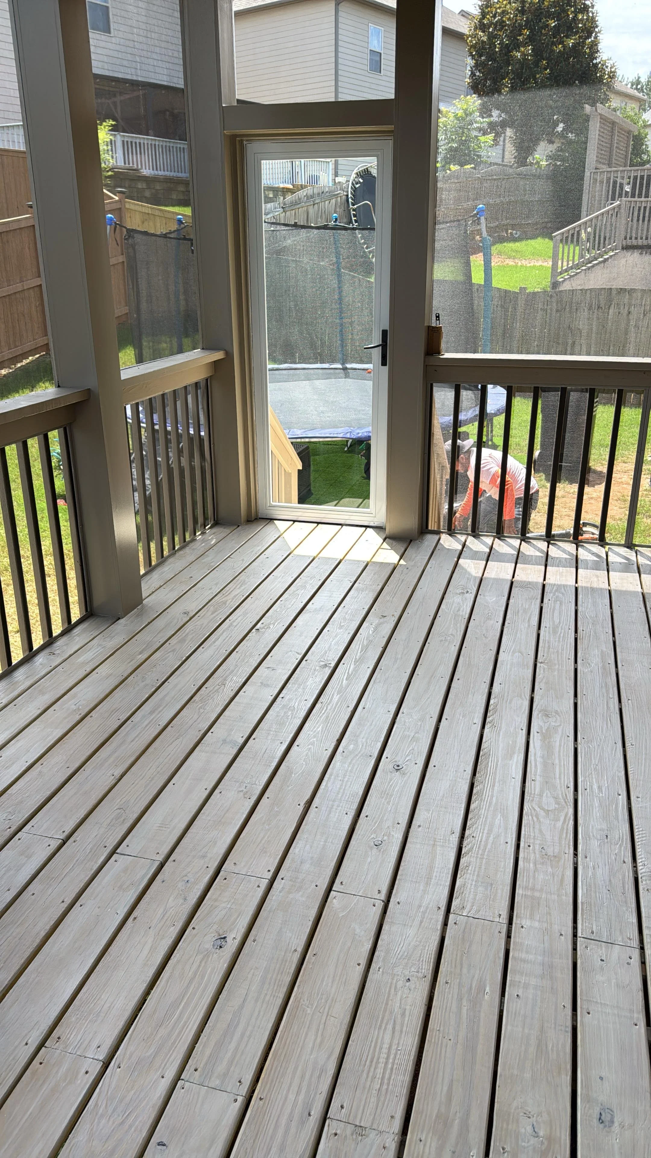 Sunlit wooden backyard deck with screened railings, a glass door, neighboring homes, lawn, and trampoline outside