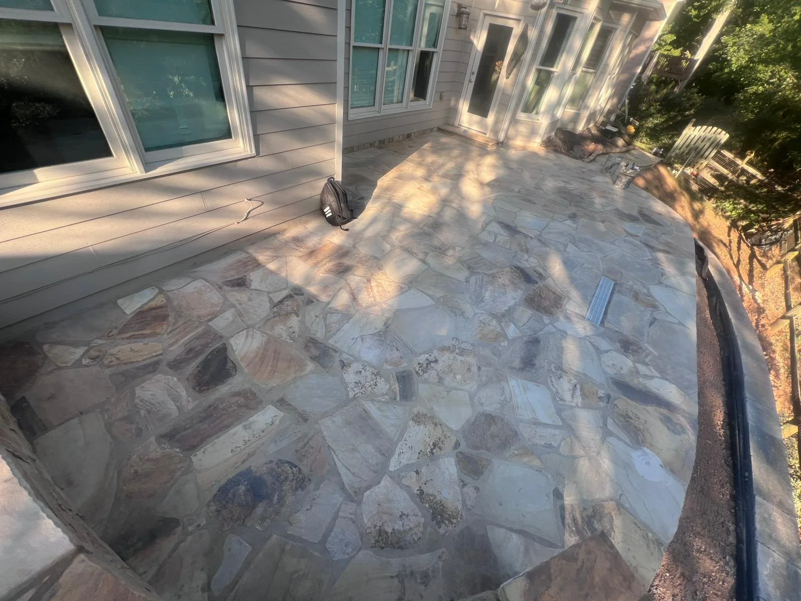 Flagstone patio beside a house with windows, doors, a backpack, outdoor chairs, and a drainage grate
