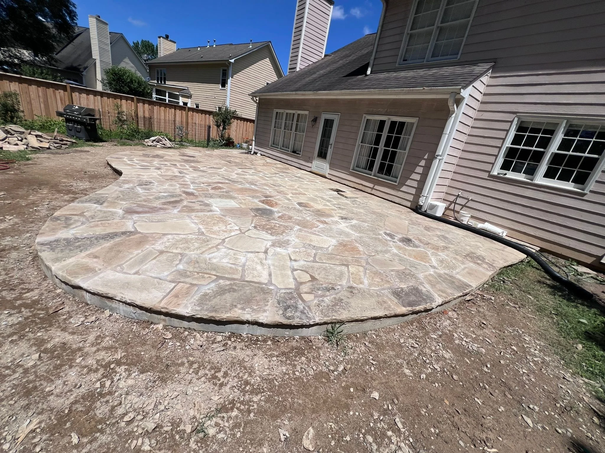 Large irregular flagstone patio beside a suburban house, with a barbecue grill, wood fence, and neighboring homes.