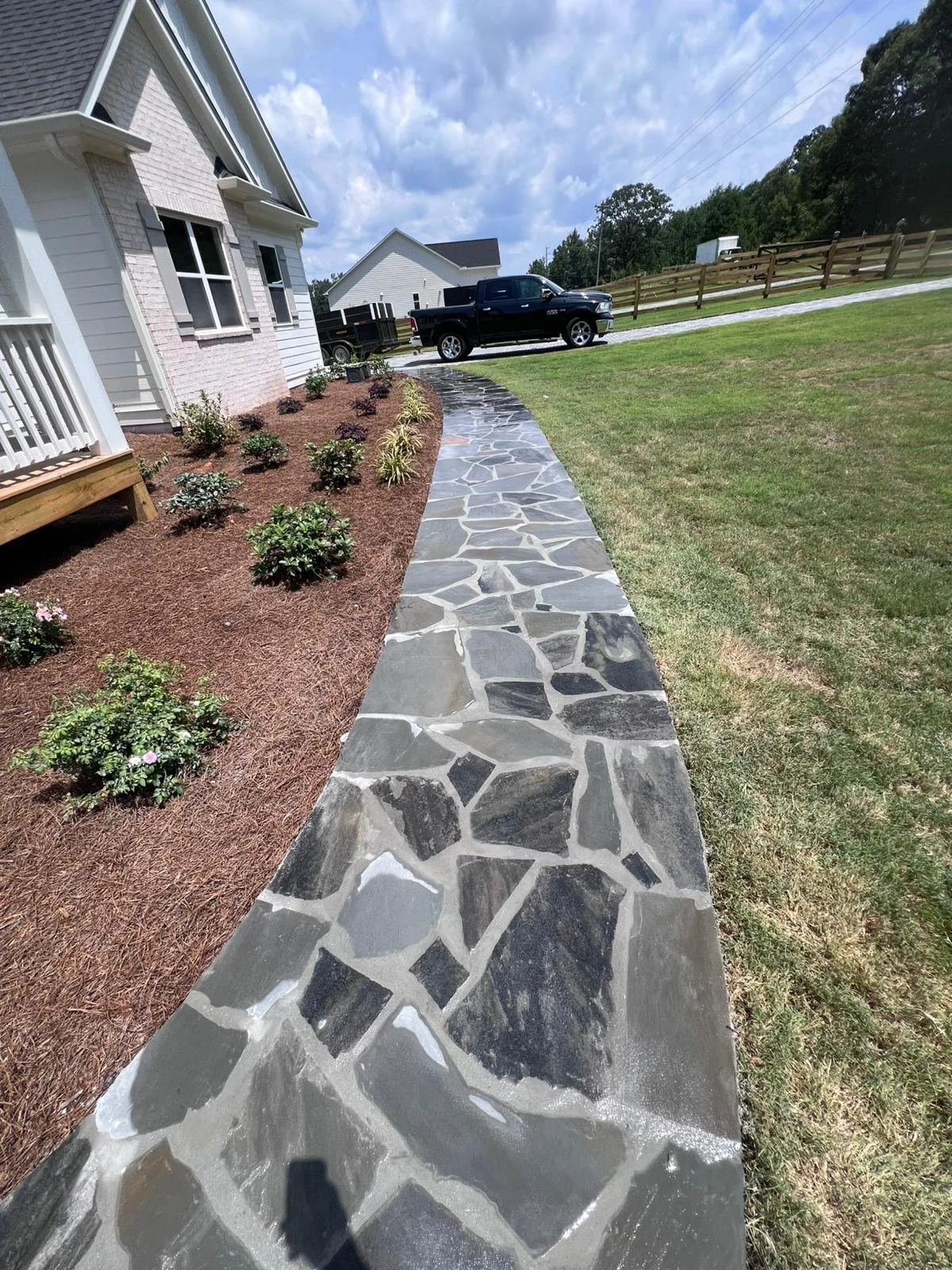 Flagstone walkway leading to a white house with landscaped flower beds, lawn, wooden fence, and black pickup truck.