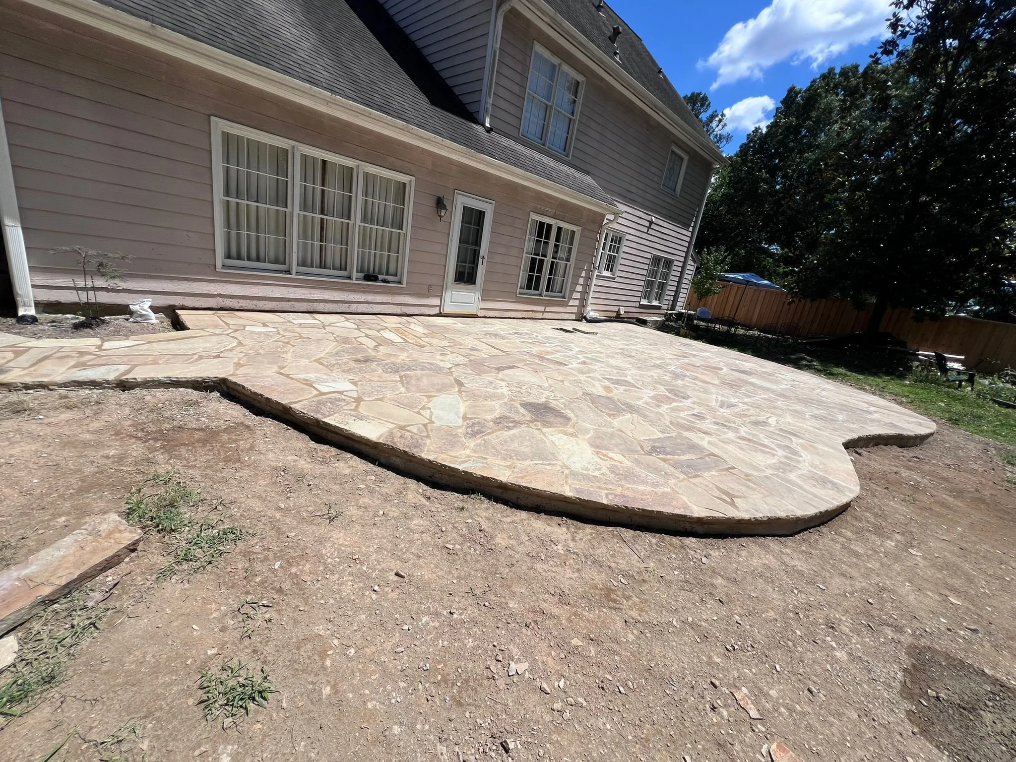 Large flagstone patio beside a beige two-story house with white-trimmed windows, trees, and a wooden fence.