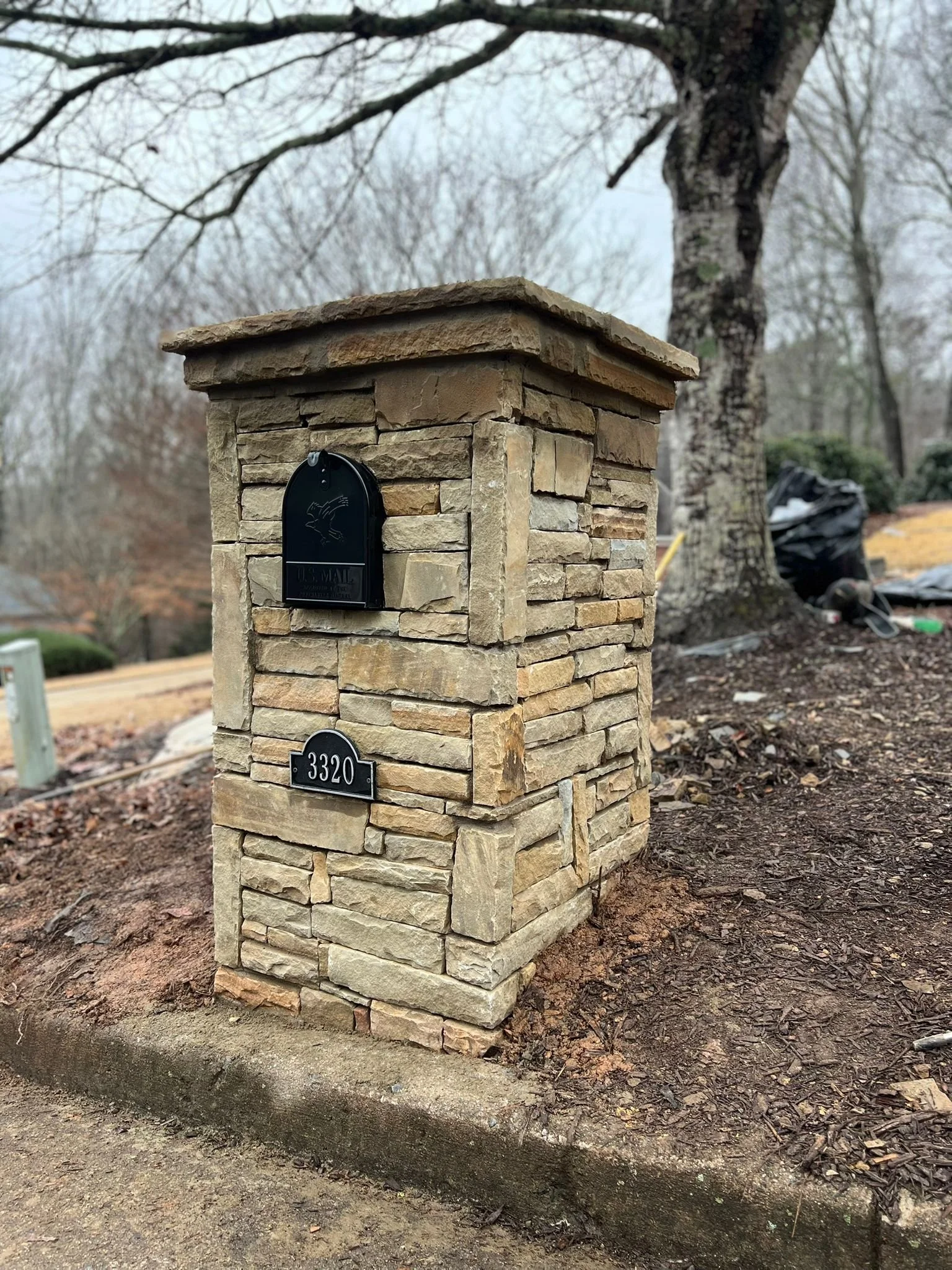 Stone mailbox pillar with black mailbox and address plaque reading 3320 in a wooded yard
