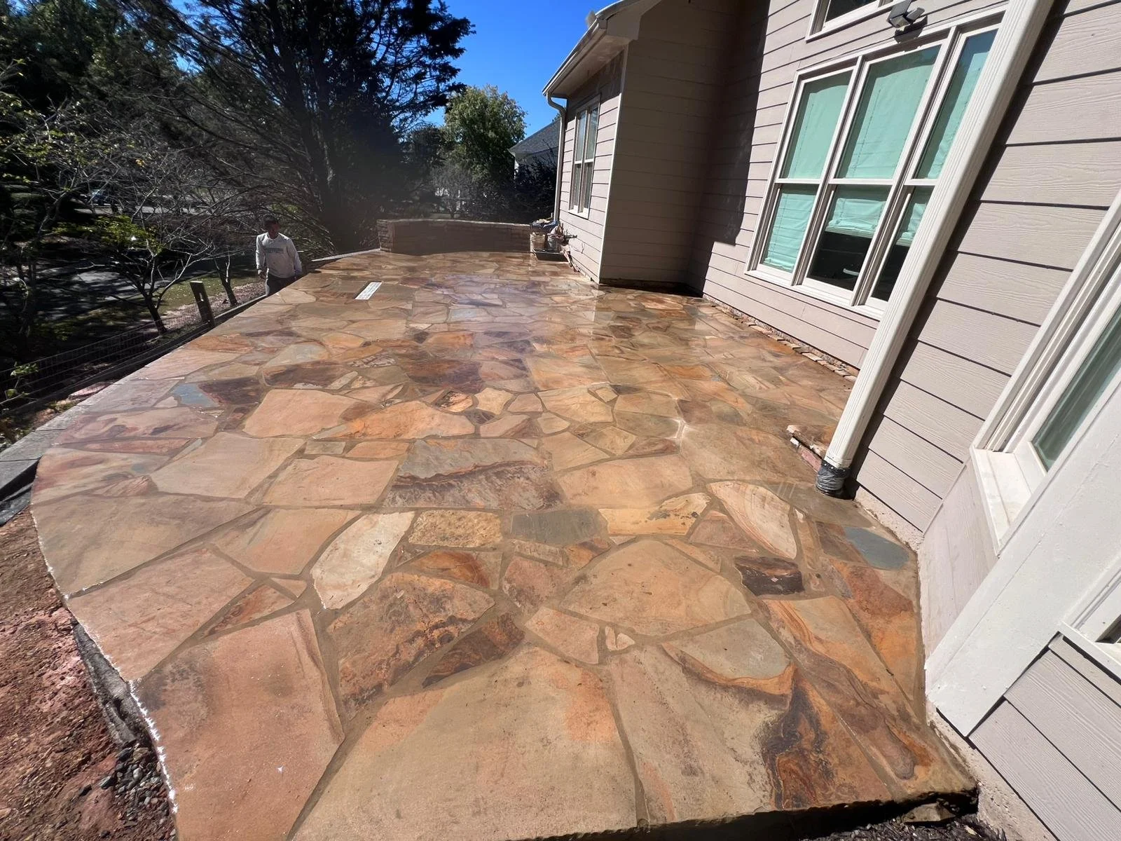 Flagstone patio beside a house with large windows, trees, and a person outdoors.